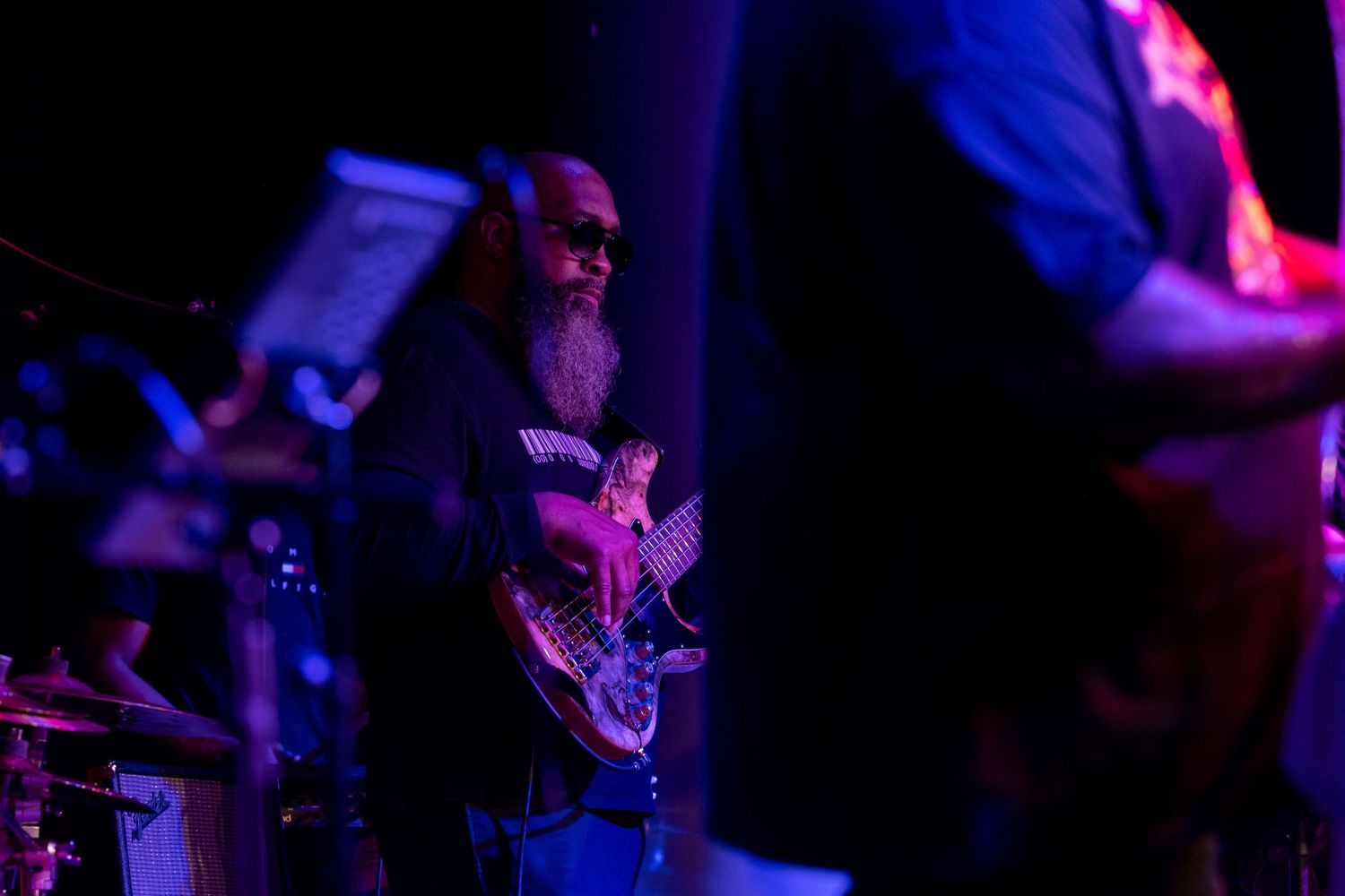 A musician plays bass guitar on a dimly lit stage illuminated by purple and blue lighting effects.