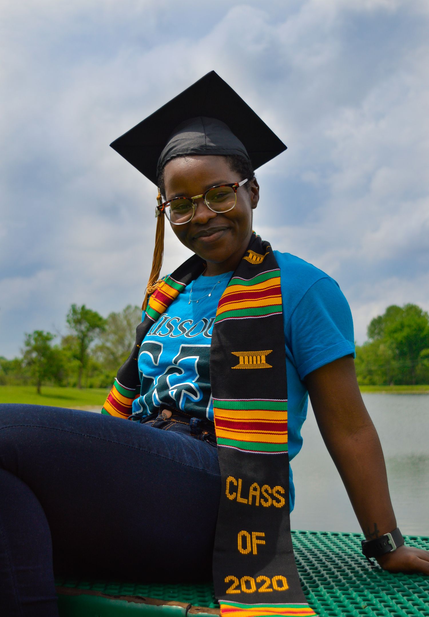 Graduate wearing Class of 2020 stole and cap poses by lake with colorful kente cloth.