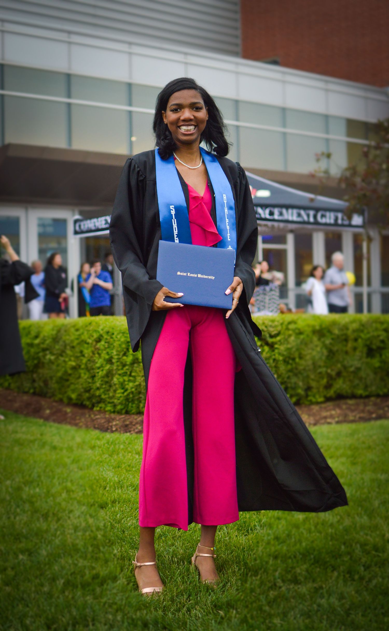 Graduate in black gown and pink skirt poses with diploma on campus lawn.