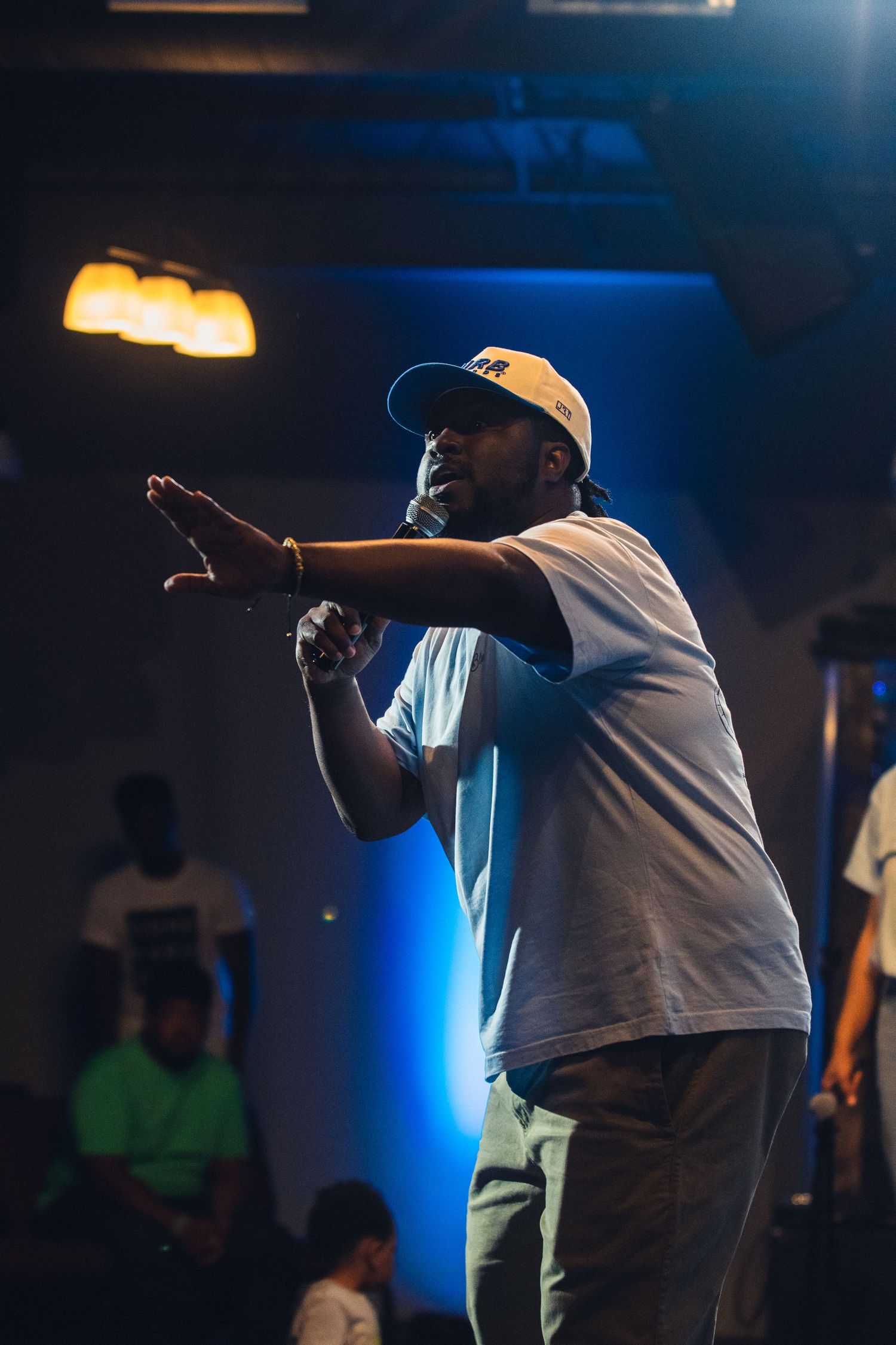 A performer in a white t-shirt and baseball cap gestures on stage with dramatic blue lighting in the background.