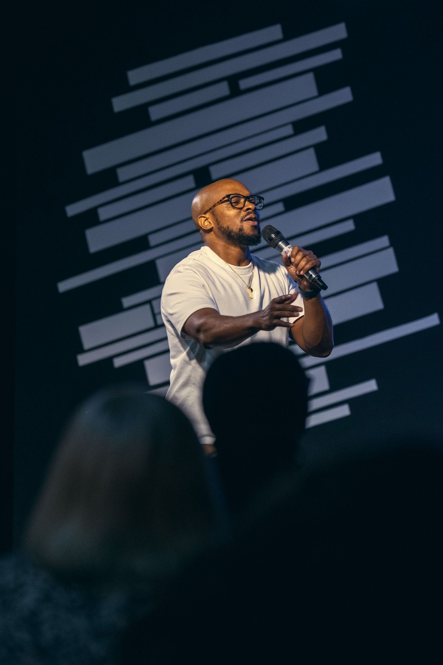 Performer in white t-shirt sings passionately on dark stage with dramatic lighting and geometric patterns in background.