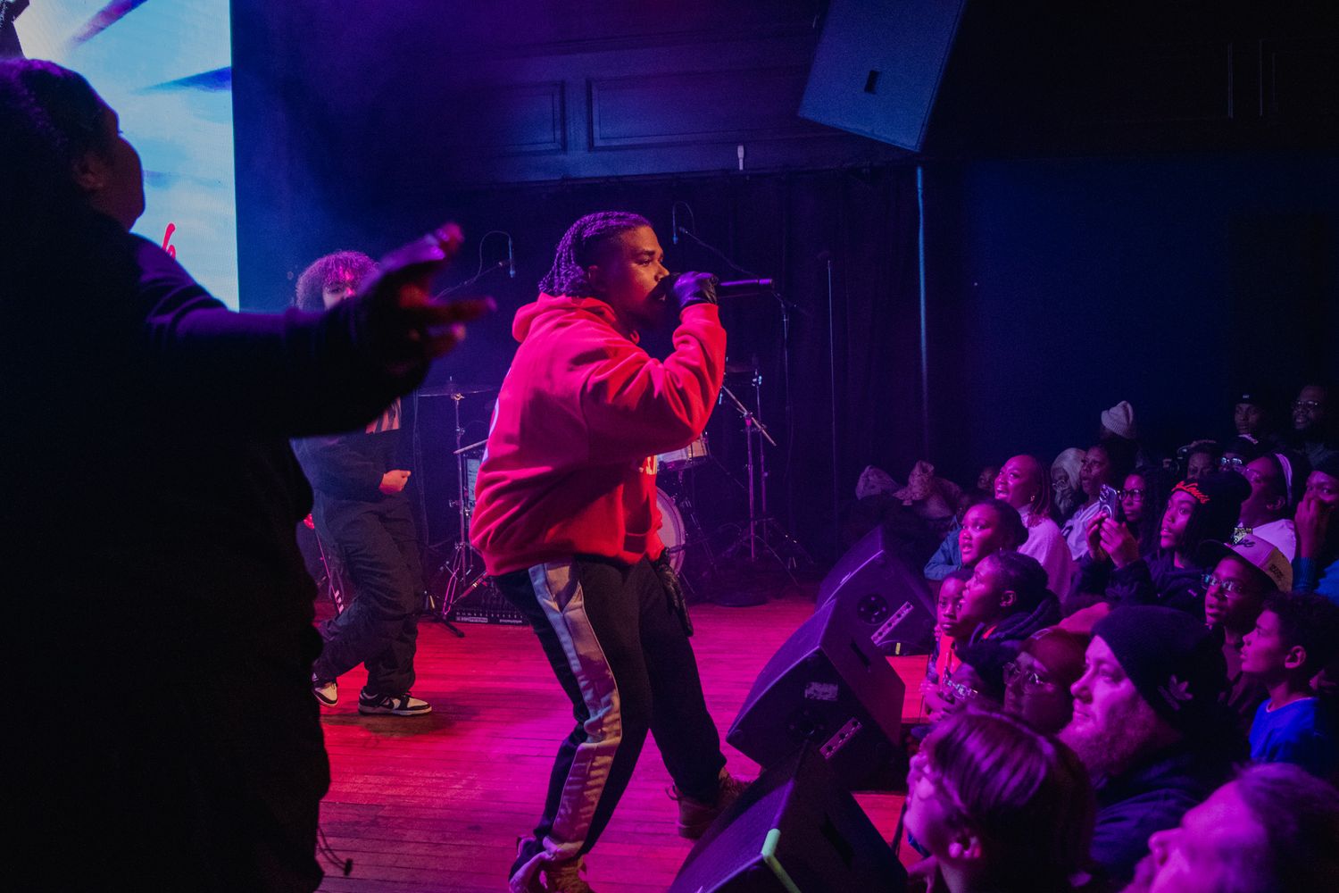 Performers dance energetically on a stage illuminated by vibrant purple and red lighting while an audience watches.