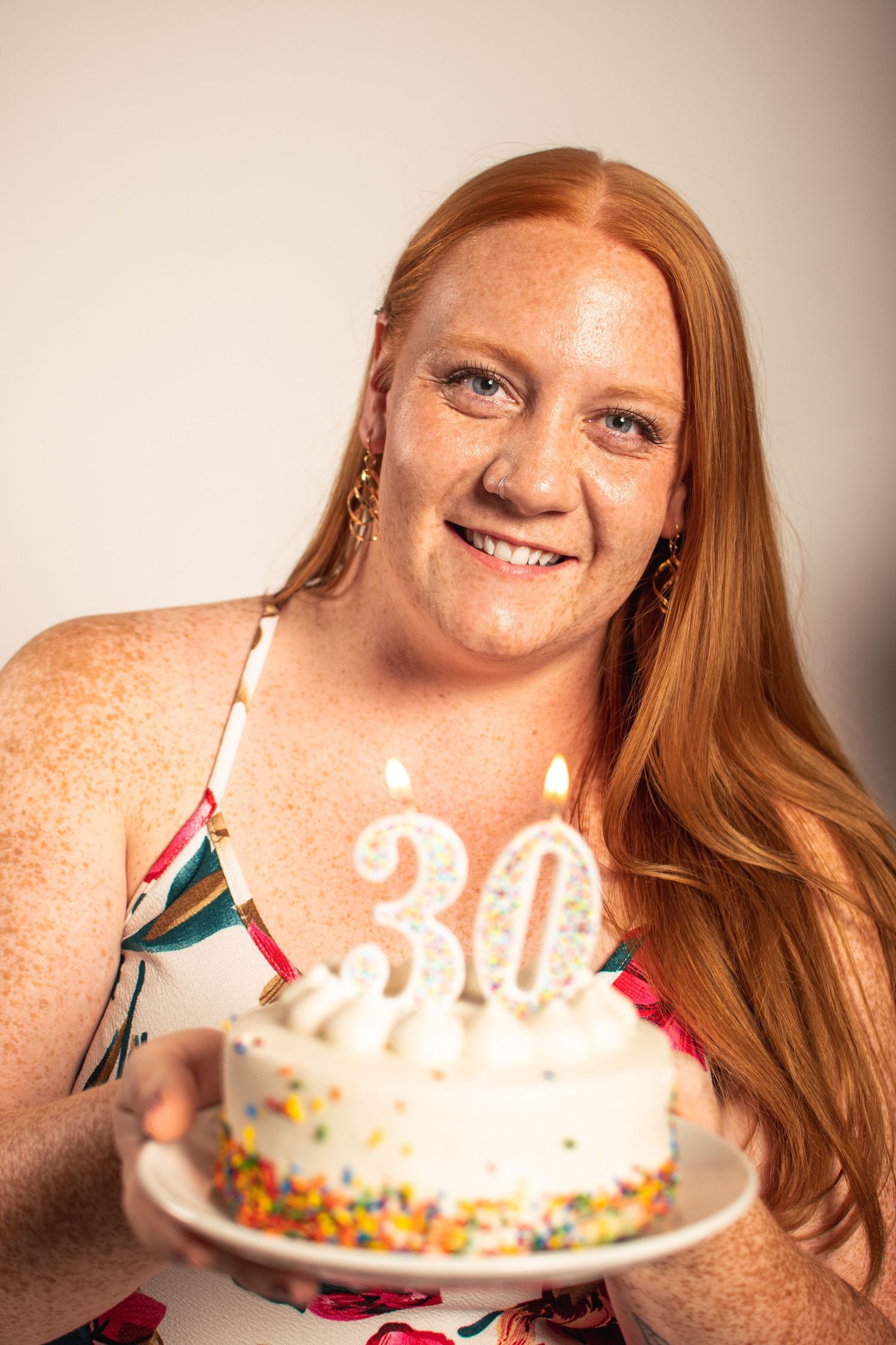 Person with long red hair holding a white birthday cake with lit candles celebrating their 30th birthday.