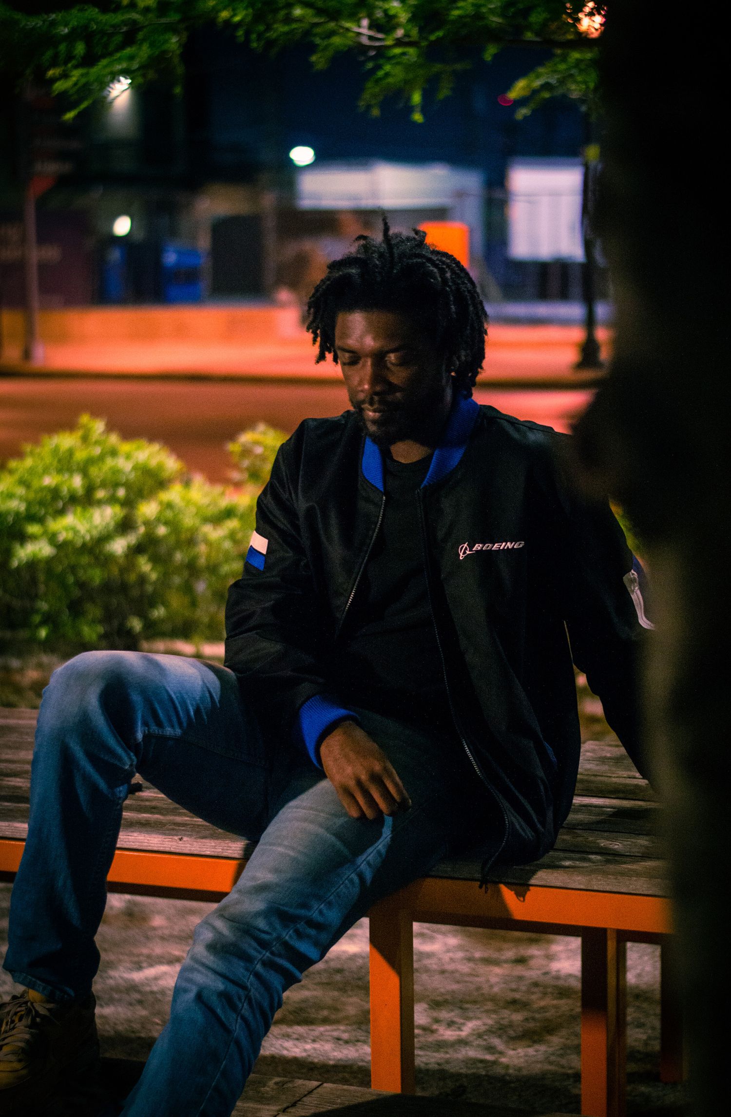 A black man sitting on wooden bench at night with moody lighting and urban background.
