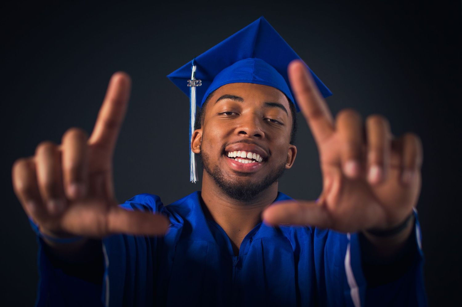 Graduate in blue cap and gown makes gesture toward camera against dark background.