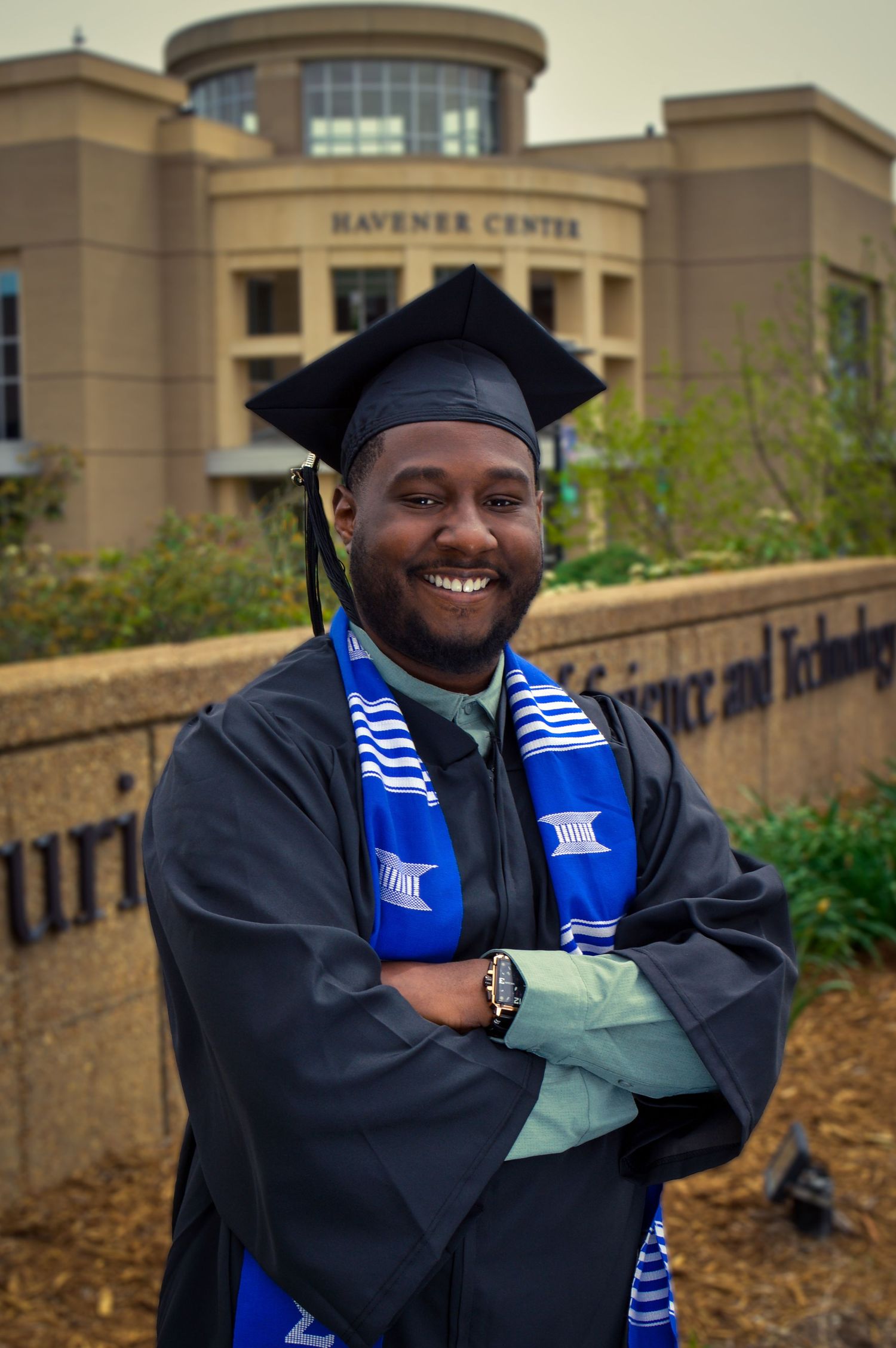 Graduate in black gown and blue stole poses confidently outside campus building.