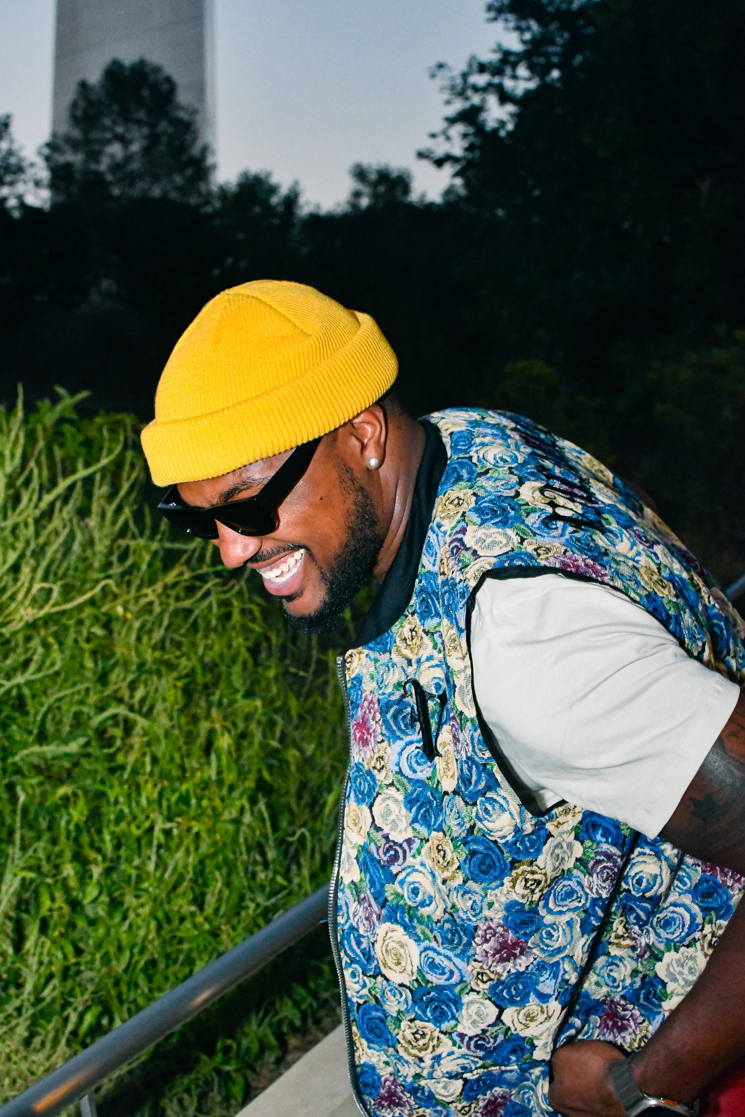 Person in yellow cap and patterned shirt smiling while leaning on railing outdoors.