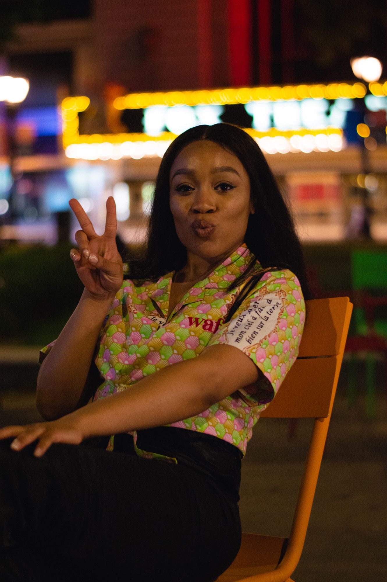 A person in a floral print top poses playfully while sitting on a wooden chair at night with city lights in background.