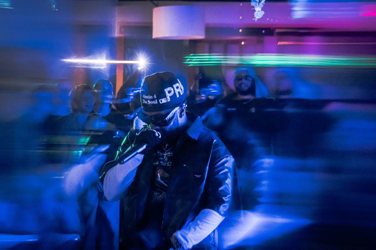 DJ performing under neon blue and purple lights in a dark club, wearing a cap and vest.