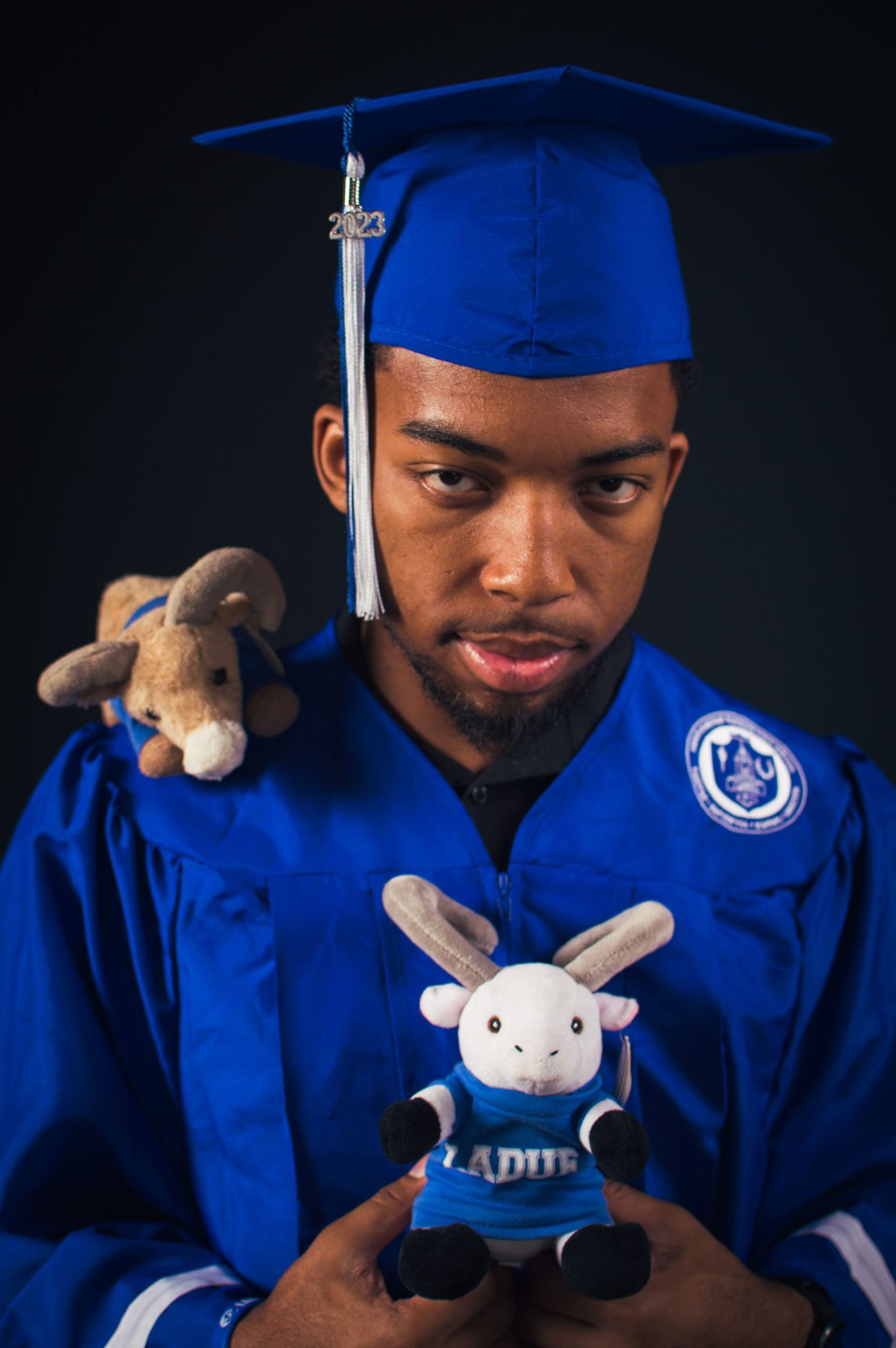 Graduate in blue cap and gown poses with small plush toy in studio portraits against dark background.