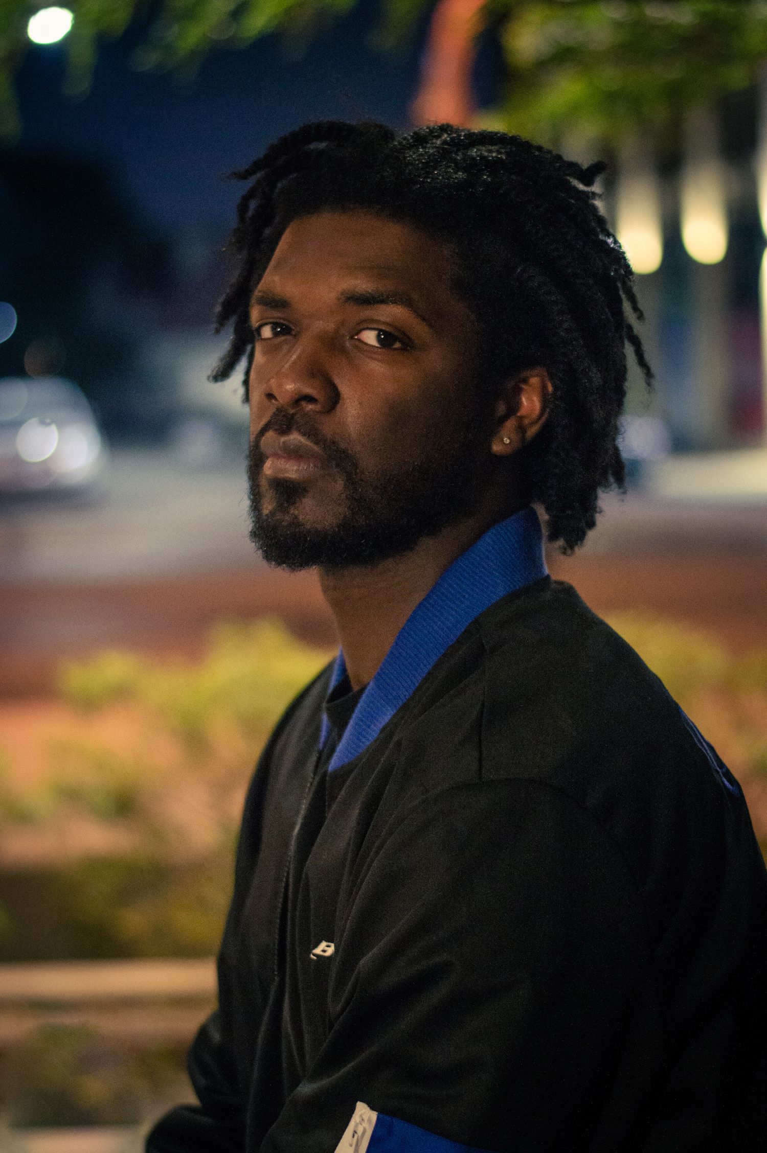 A black man in a black jacket and blue shirt shown in a nighttime outdoor setting with atmospheric lighting.