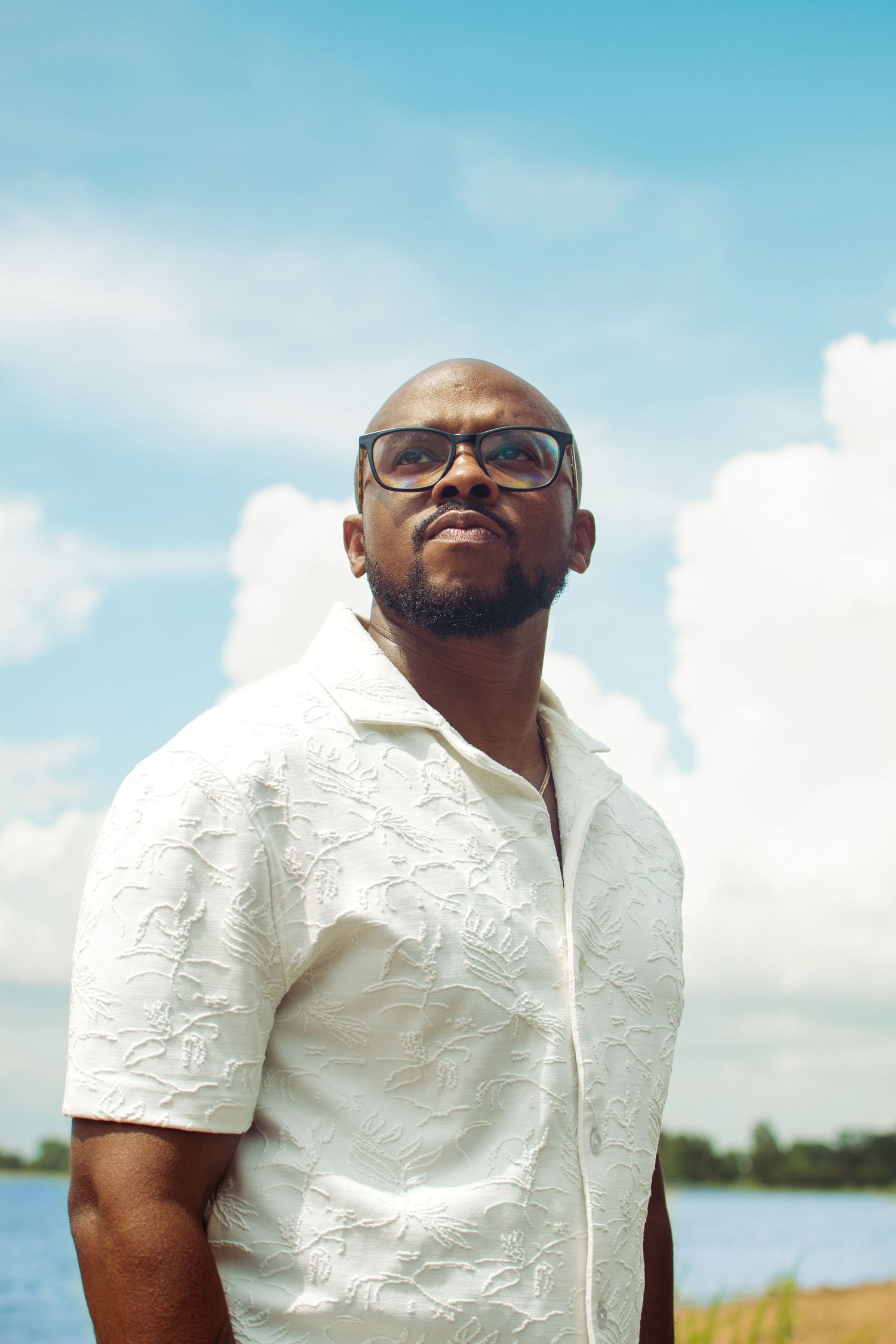 A person wearing glasses and a white embroidered shirt smiles joyfully against a bright blue sky and water backdrop.