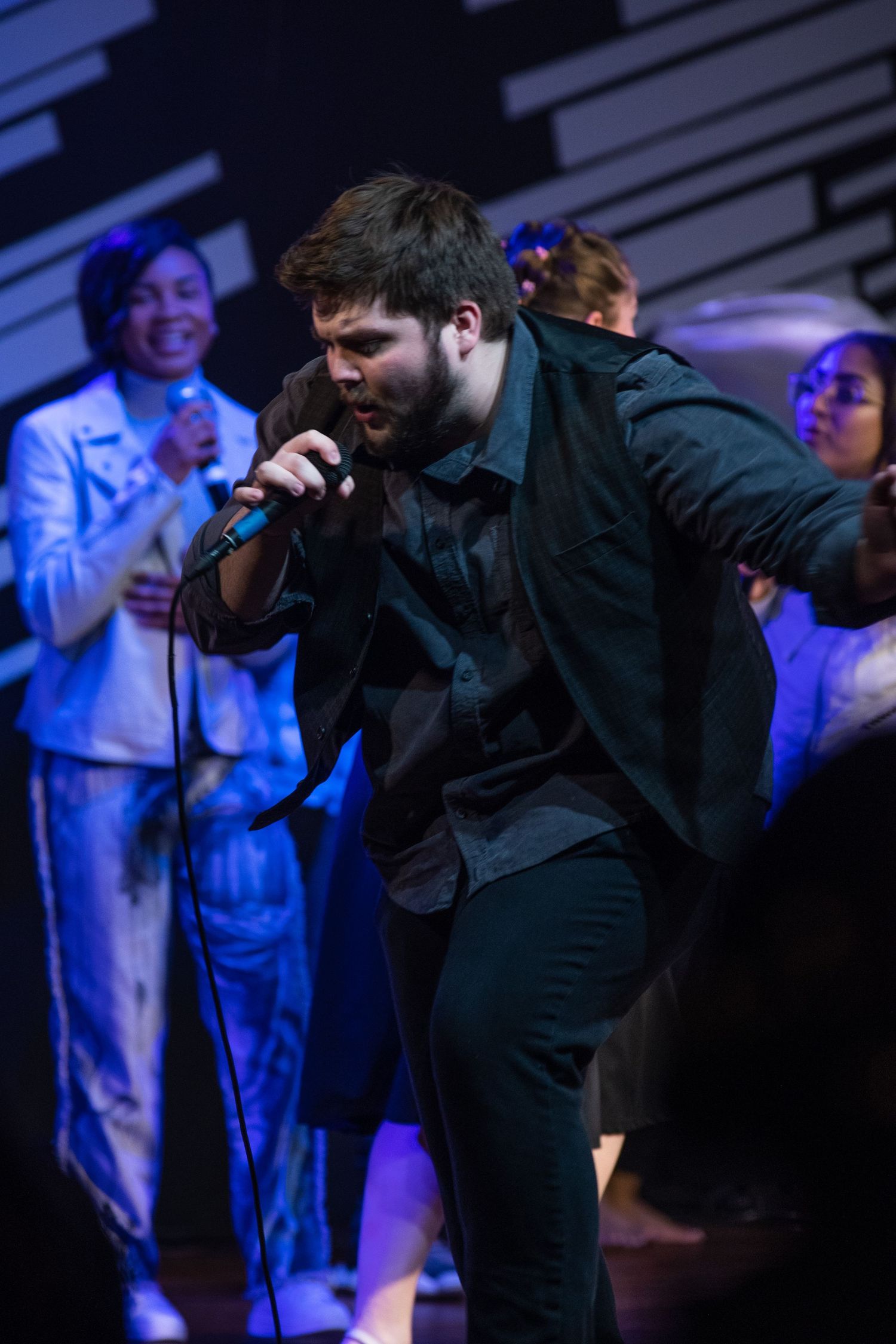 A performer sings passionately on stage with dramatic blue lighting while others stand in the background.