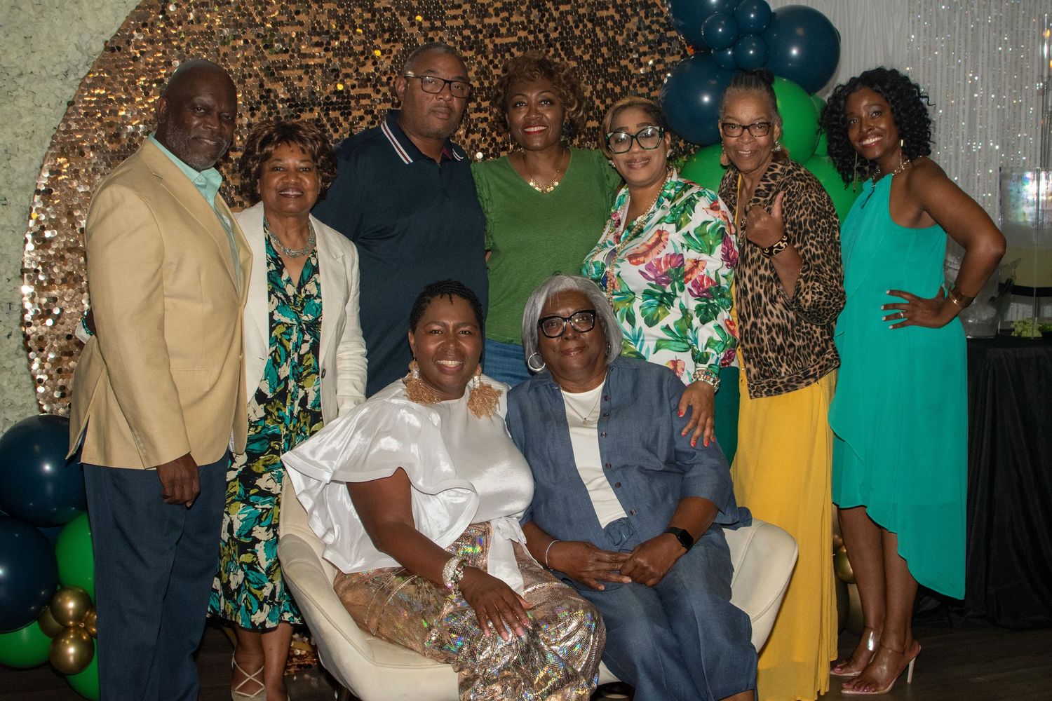 A group in coordinated green and white outfits poses together at a celebration with balloons and decorative backdrop.