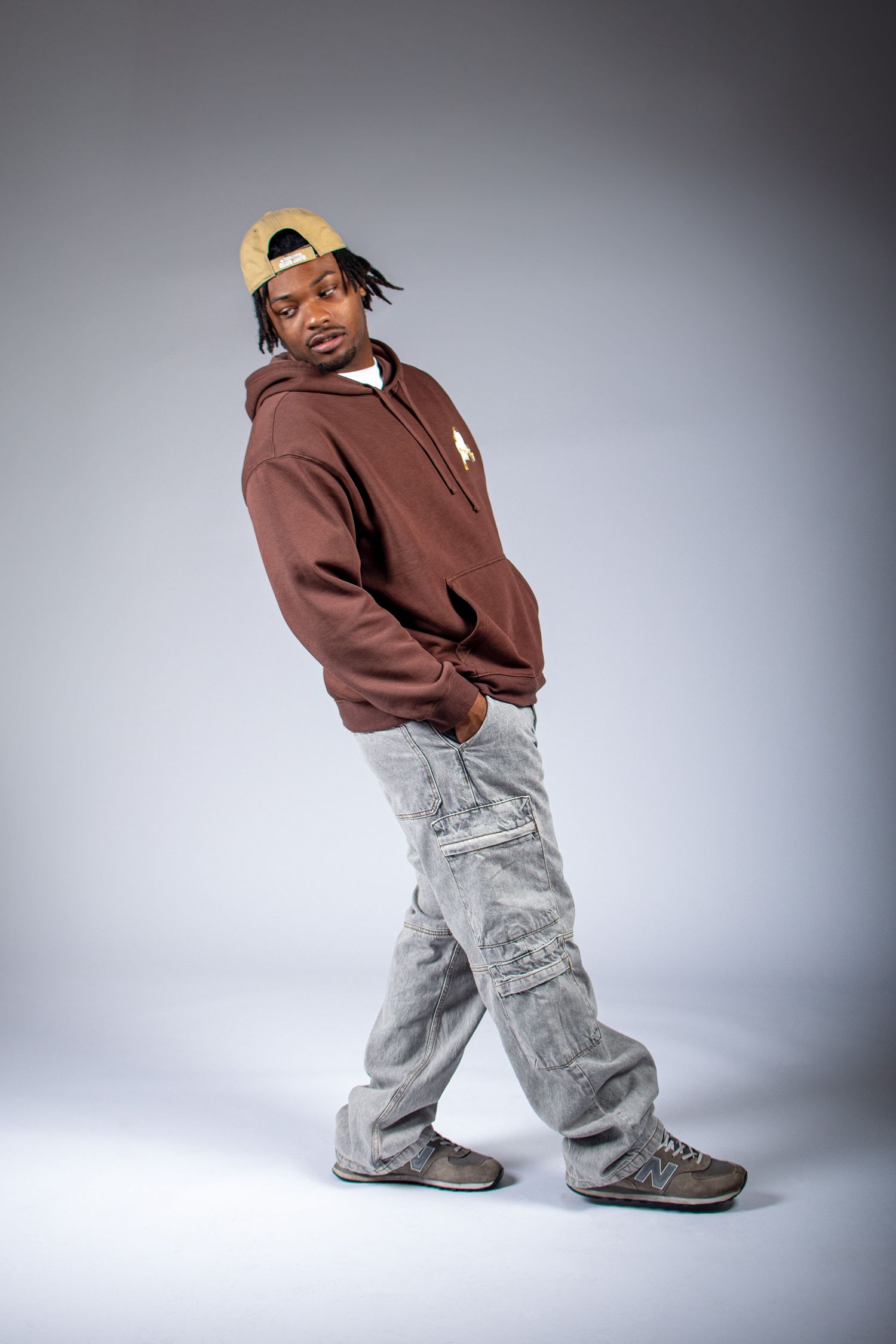 A figure in a brown sweater and light gray cargo pants strikes dynamic dance poses against a gray studio backdrop.