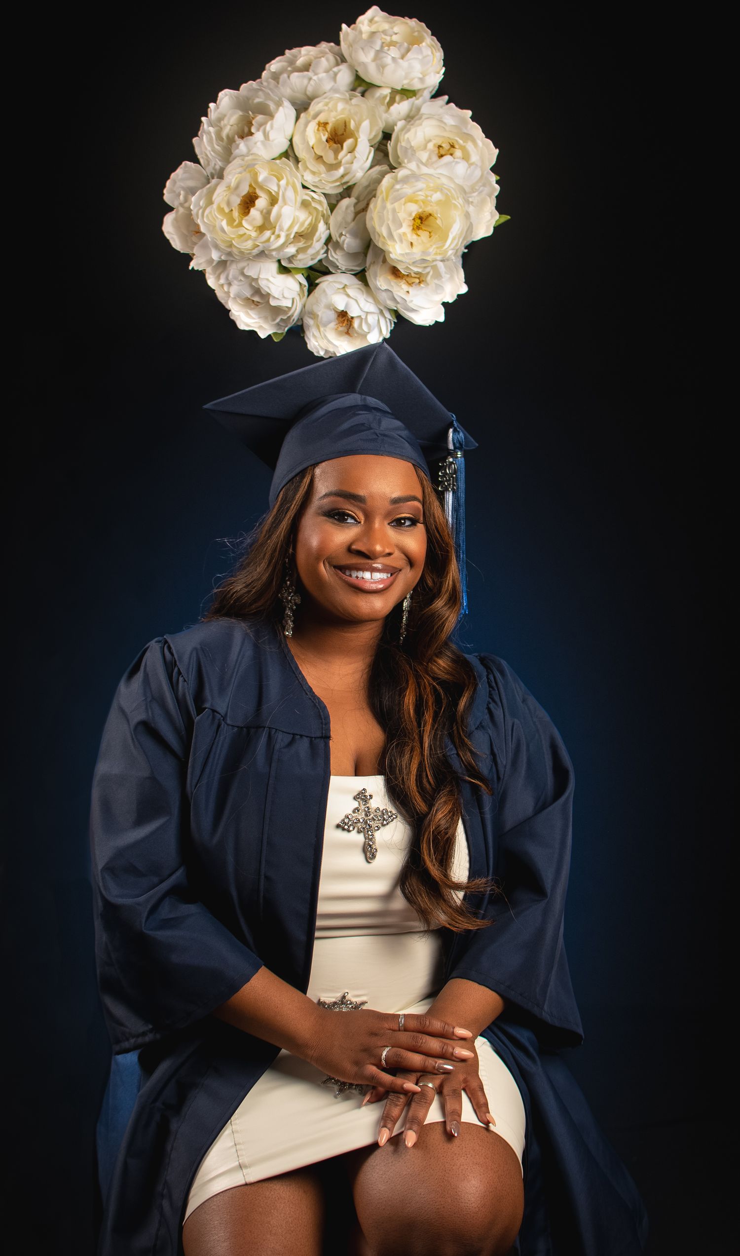 Graduate in navy cap and gown poses with white roses against dark background for professional photos.