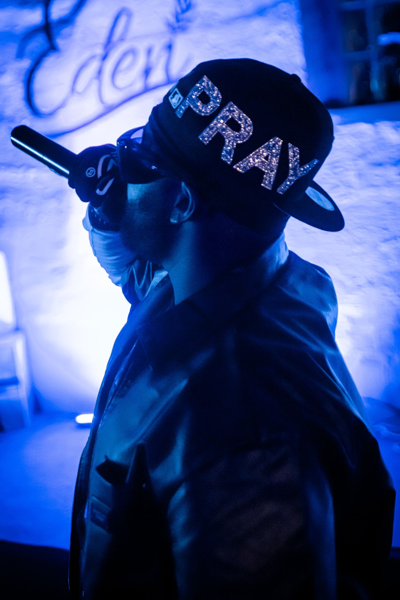 Performer in blue light wearing a sparkly PRAY snapback cap holding a microphone at Eden venue.