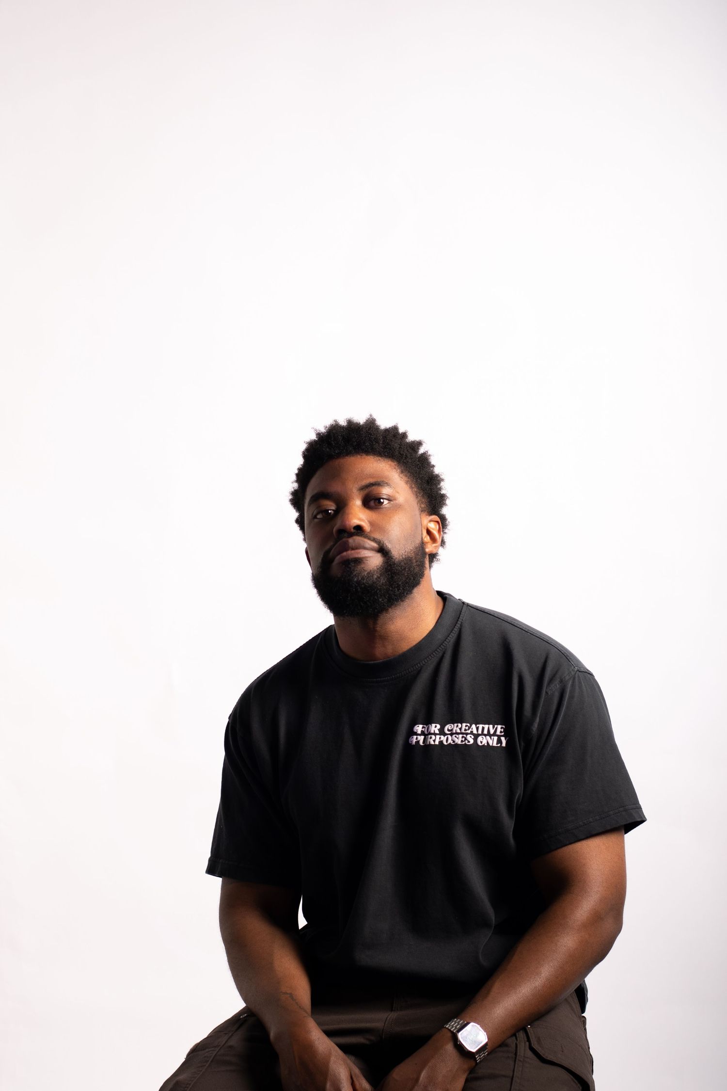 A bearded Black man sits confidently in a black graphic t-shirt against a white background.