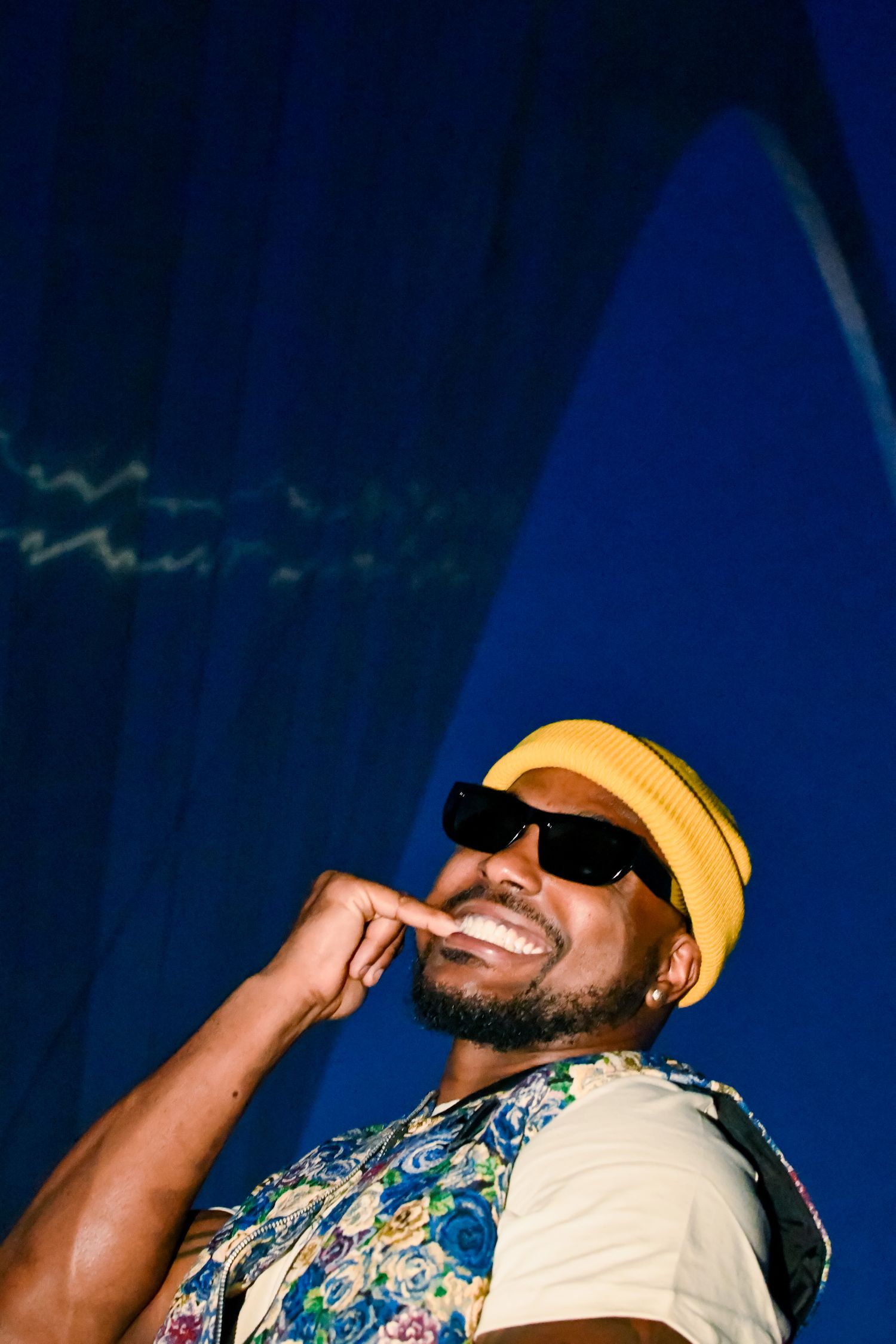Person wearing sunglasses and yellow cap smiles while wearing floral print shirt against dark blue background.