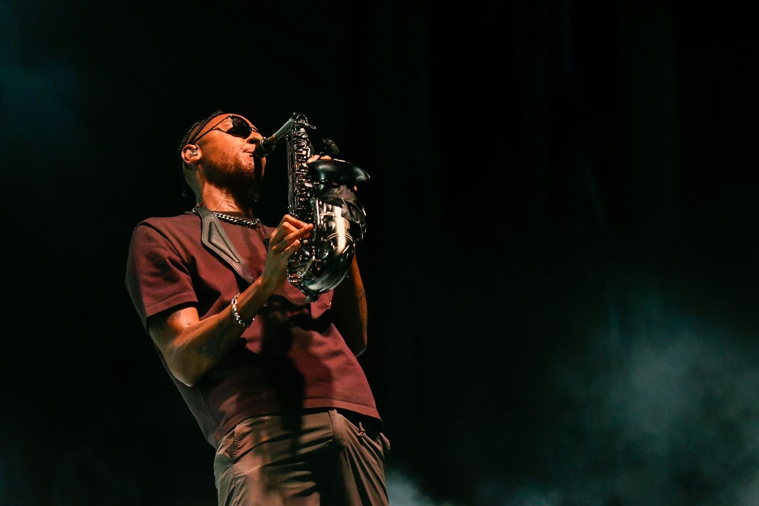 Musician performs passionately on stage wearing a burgundy shirt while holding a microphone in a dark concert setting.
