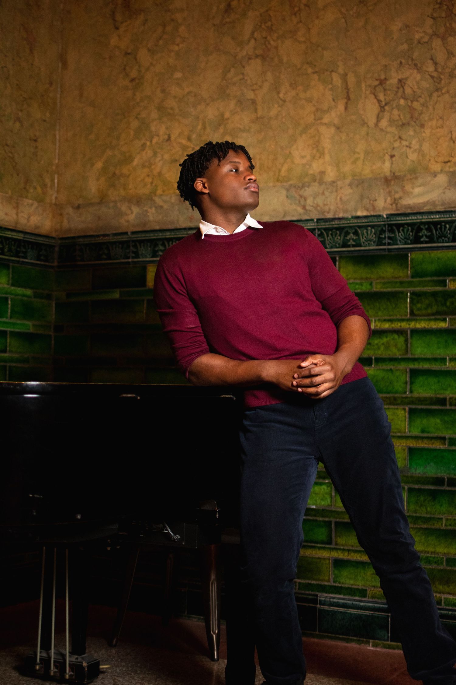 A student in a burgundy sweater and navy pants sits against a green wall in a classroom setting.