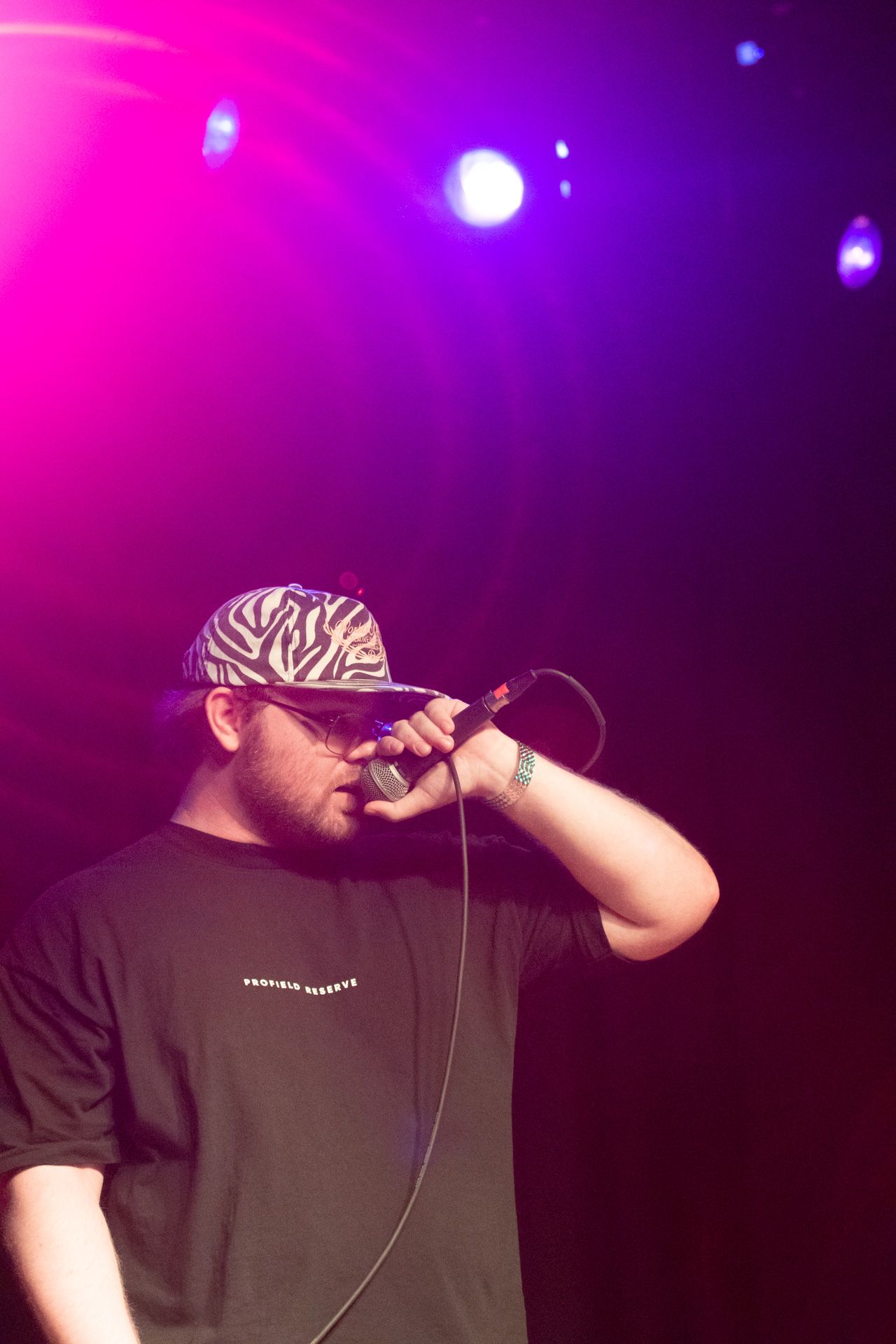 Performer wearing a black t-shirt and zebra-print hat sings into microphone on dark stage with pink and purple lighting.