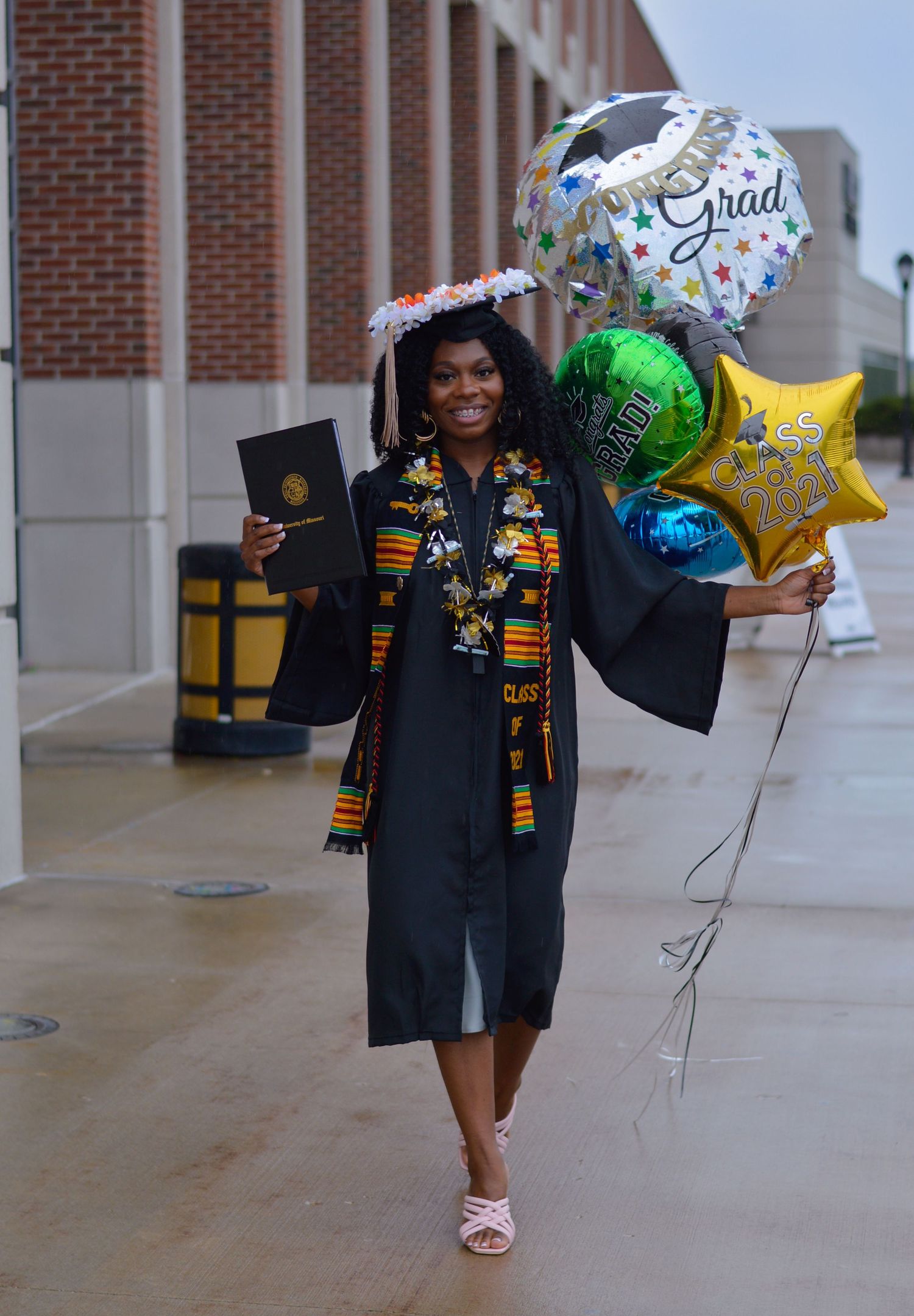 Graduates in black caps and gowns with kente stoles celebrate with balloons and joyful poses on a brick walkway.