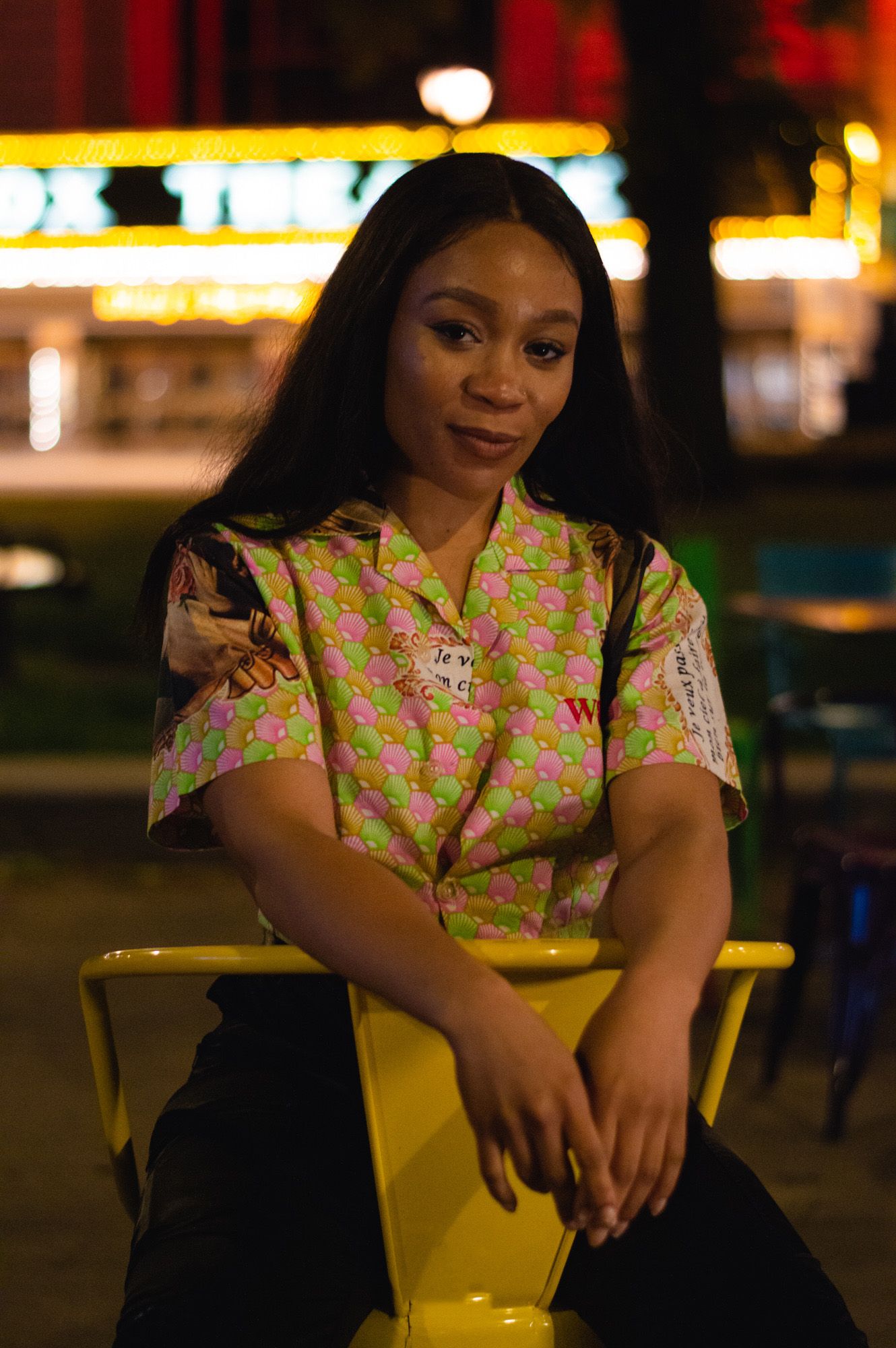 Person in floral yellow dress sits on yellow chair at night with city lights in background.