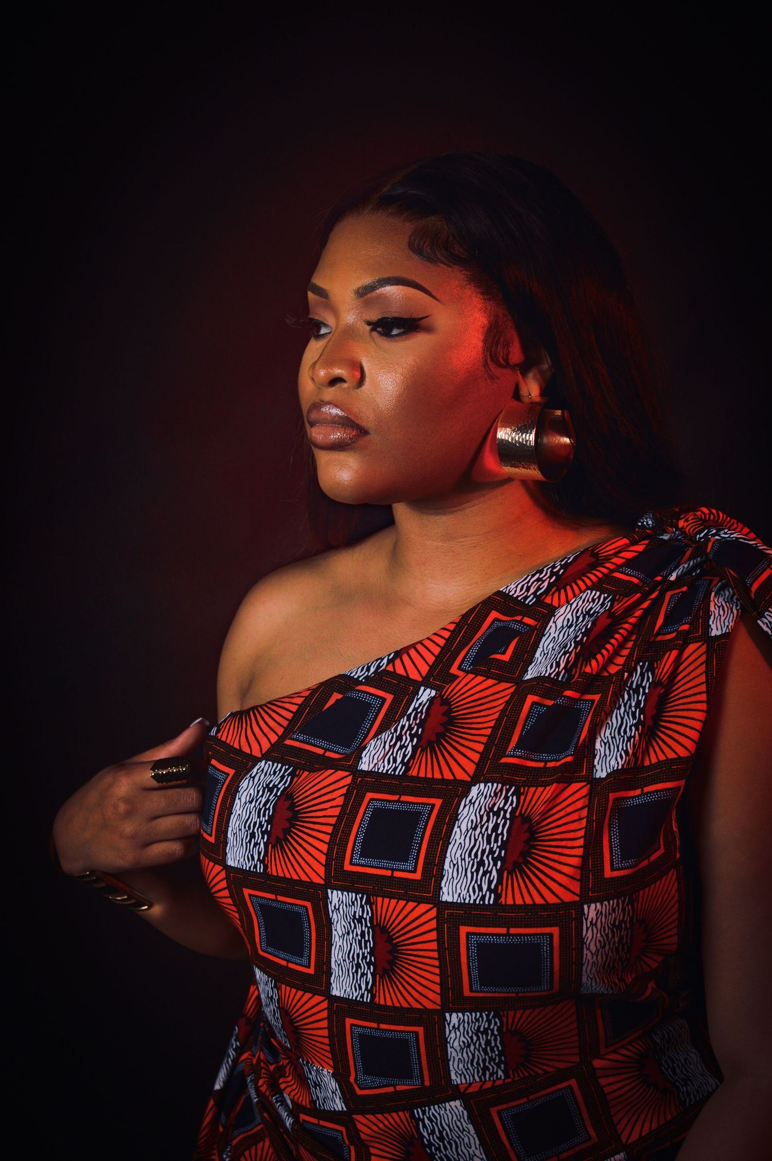 A model poses in a vibrant red and black geometric patterned one-shoulder dress against a dark background.