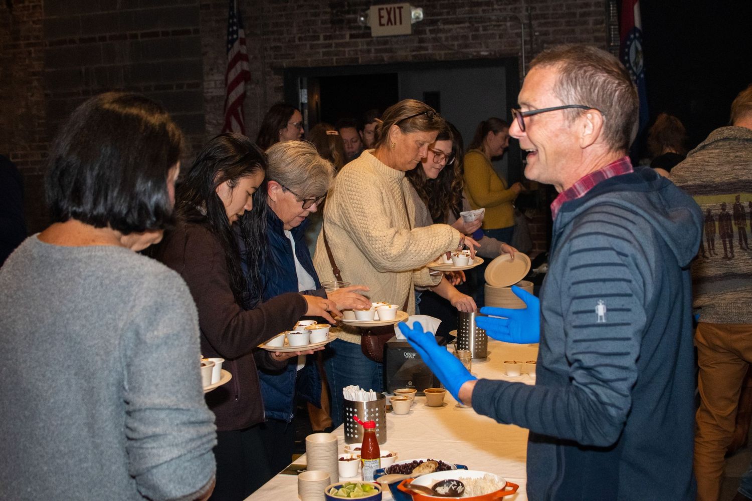A group of people gather around a table sharing food and drinks at a casual indoor event.