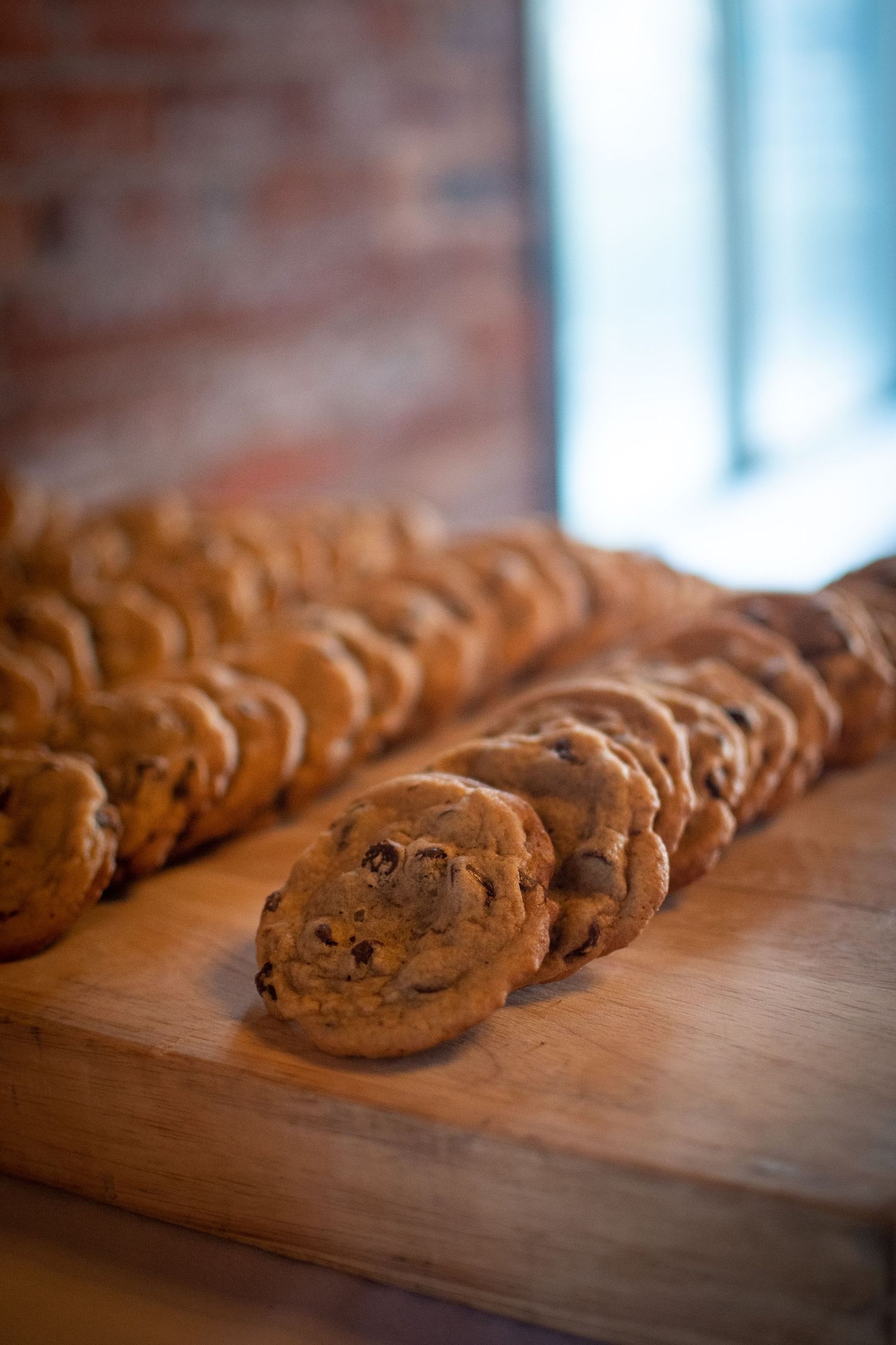 Fresh baked chocolate chip cookies lined up on a wooden surface in natural lighting.