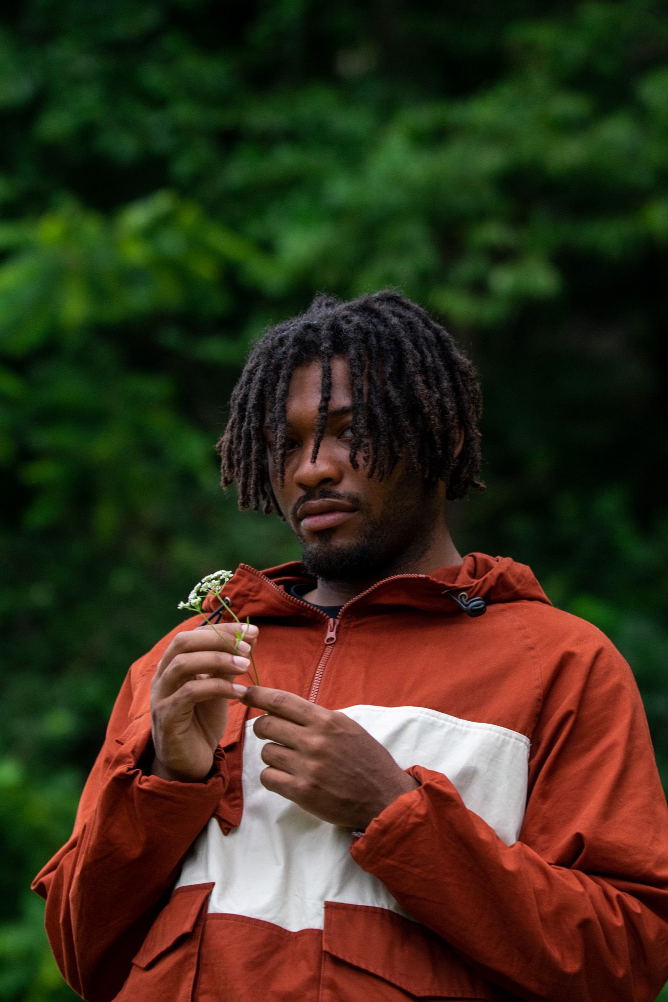 Person in a rust-colored and white windbreaker jacket stands outdoors against a blurred green forest background.