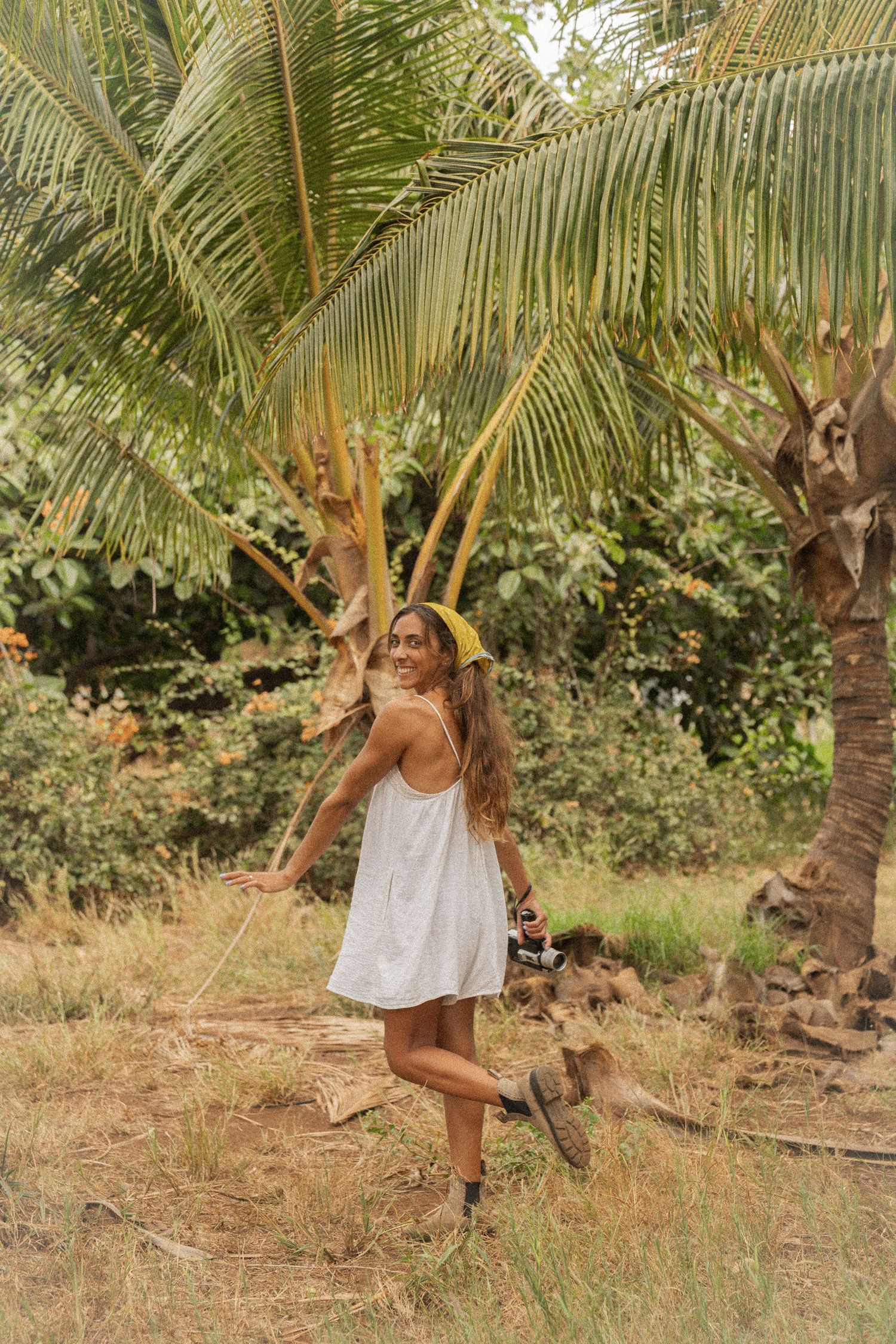 A person in a white dress dances and spins under tropical palm trees in a garden setting.
