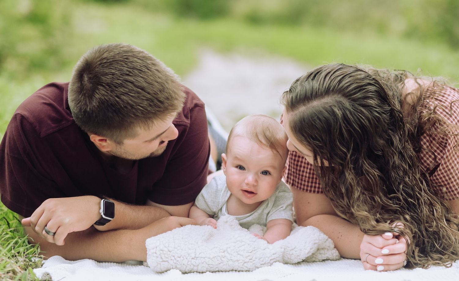 Young parents lying on a blanket outdoors smiling at their baby between them.