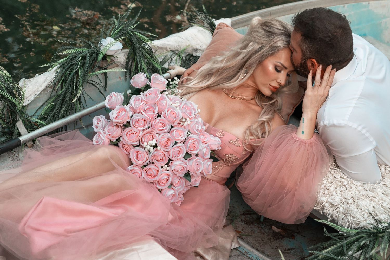 A woman in a pink tulle gown holds roses while a man leans close to her in a boat.