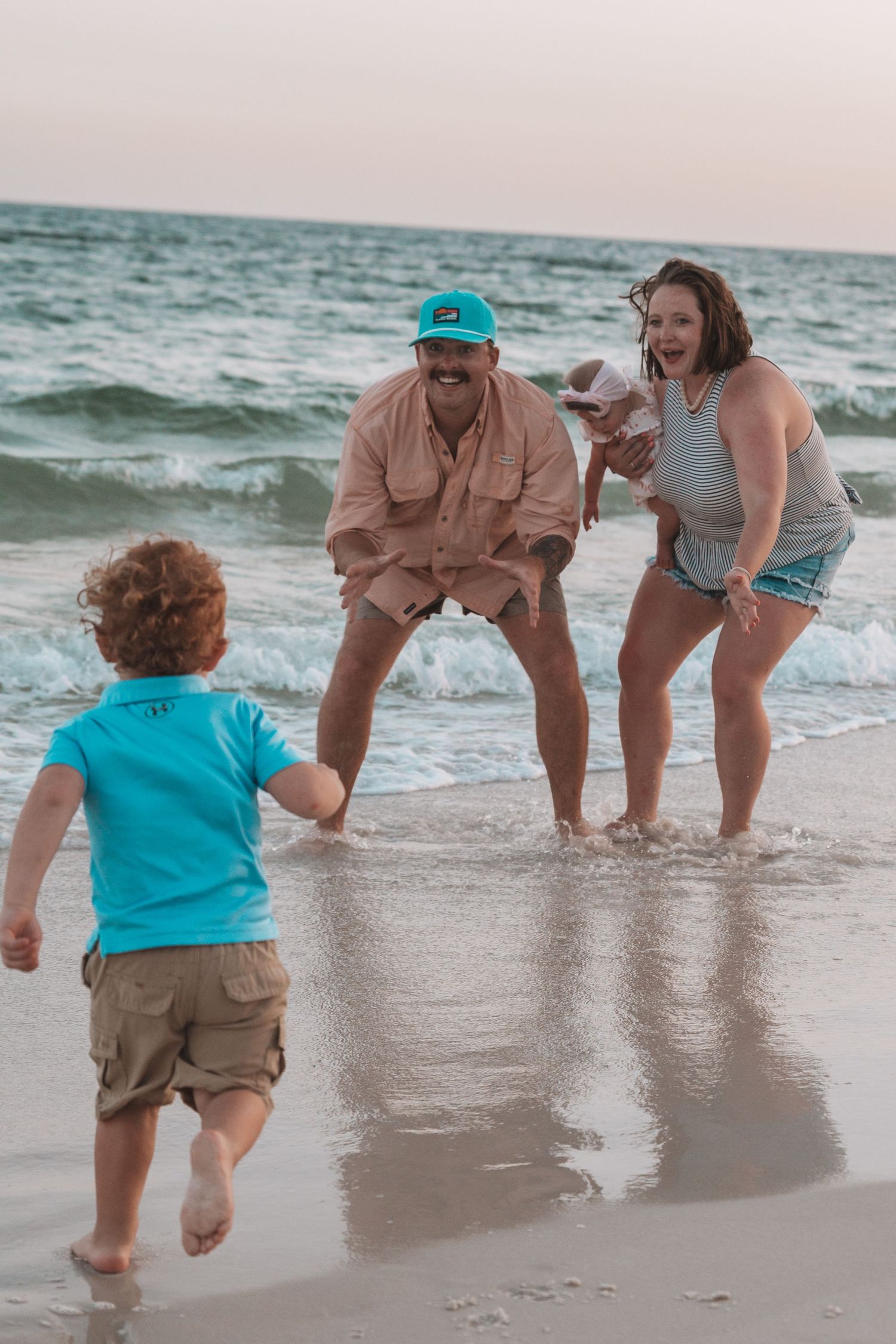 Family playing on beach at sunset, parents laughing as toddler runs toward them in shallow ocean waves.