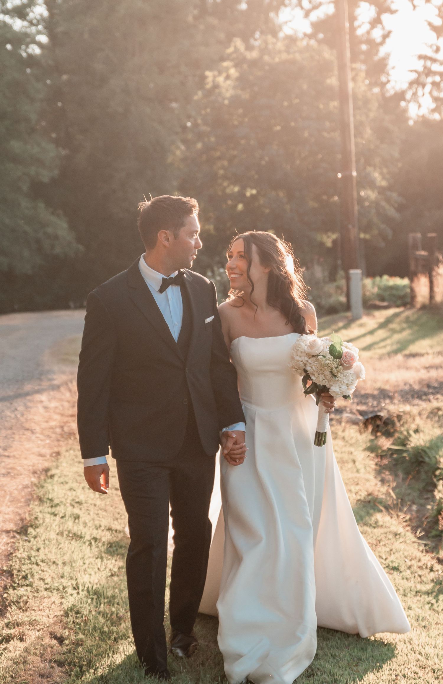 A bride and groom walk hand-in-hand outdoors in golden sunset light, she holds a white bouquet in a strapless gown.