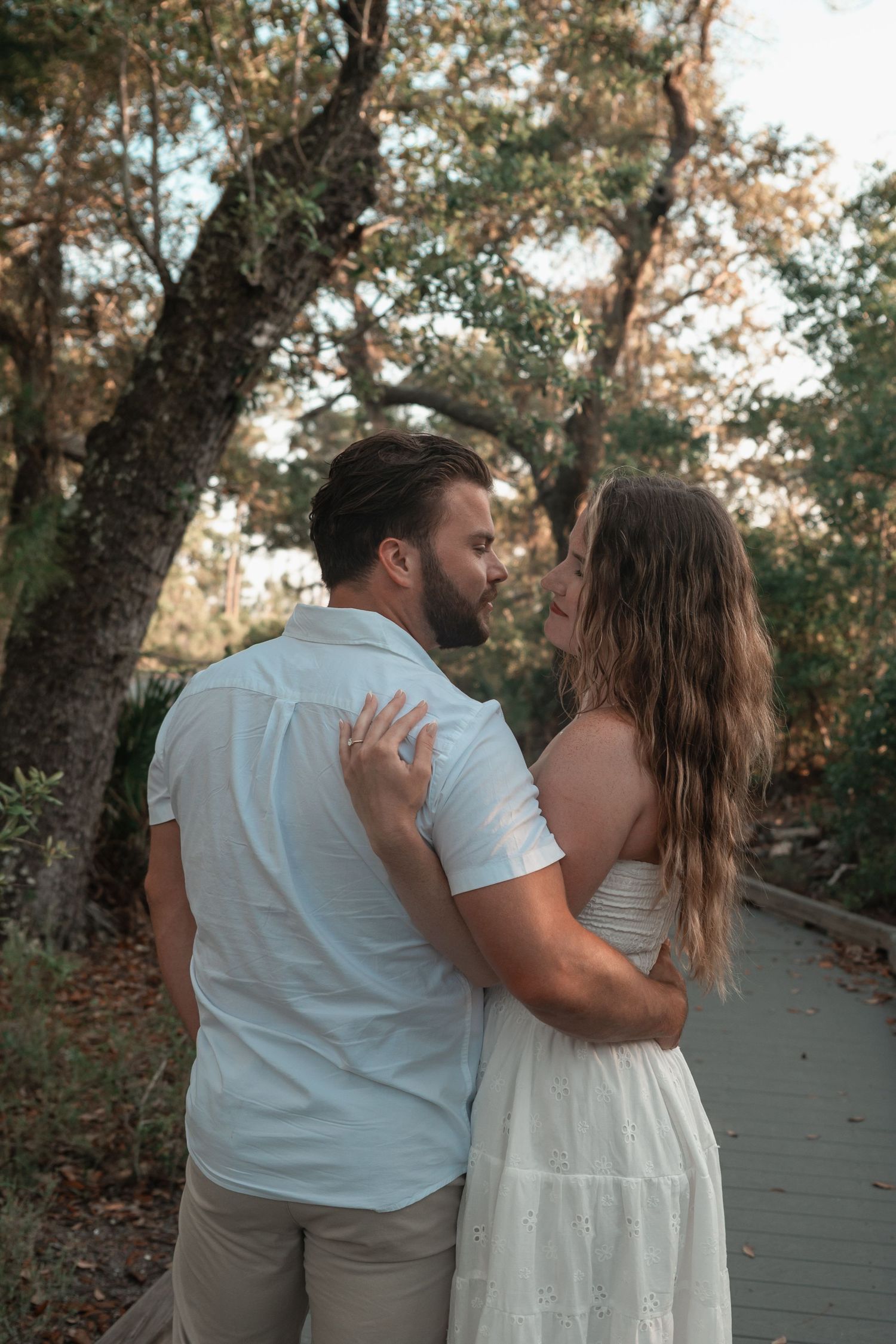 Couple sharing a kiss on a wooden boardwalk surrounded by trees, woman in white strapless dress.
