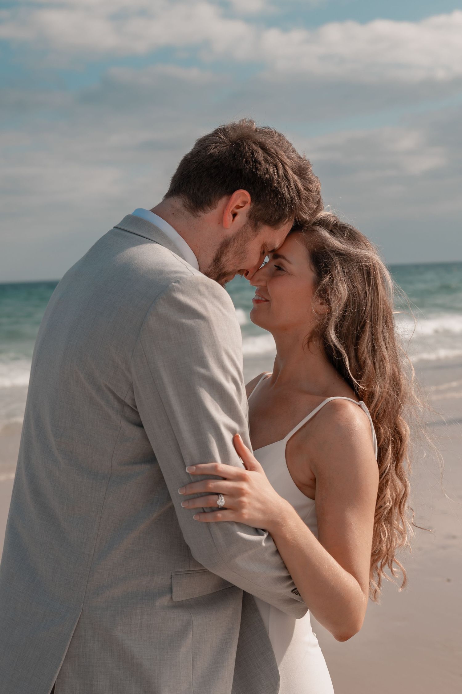 Romantic couple in wedding attire embracing on a beach with ocean waves behind them.