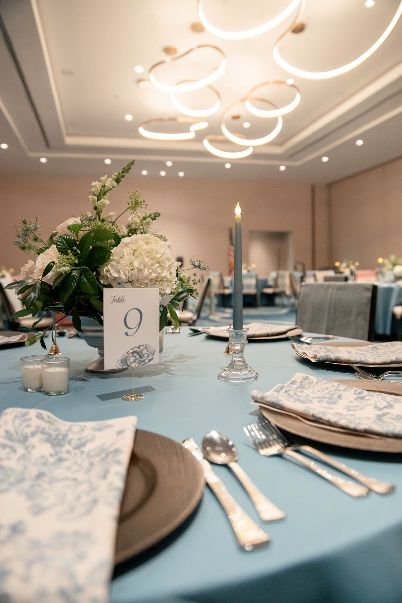 Elegant wedding reception table with blue linen, floral centerpiece, candle, and table number 9 in a ballroom.