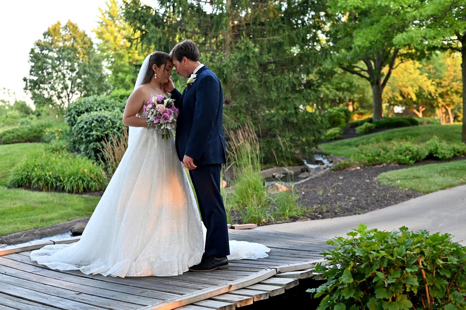 Bride and groom sharing an intimate moment on a wooden bridge surrounded by lush garden greenery at dusk.