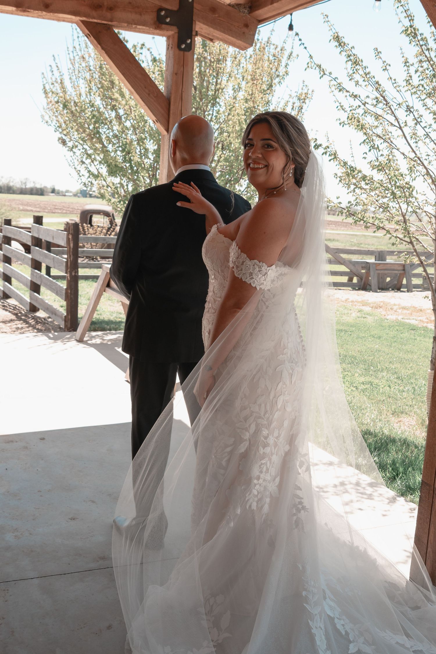 Bride in lace wedding gown and veil shares first look moment with groom under rustic wooden pergola outdoors.
