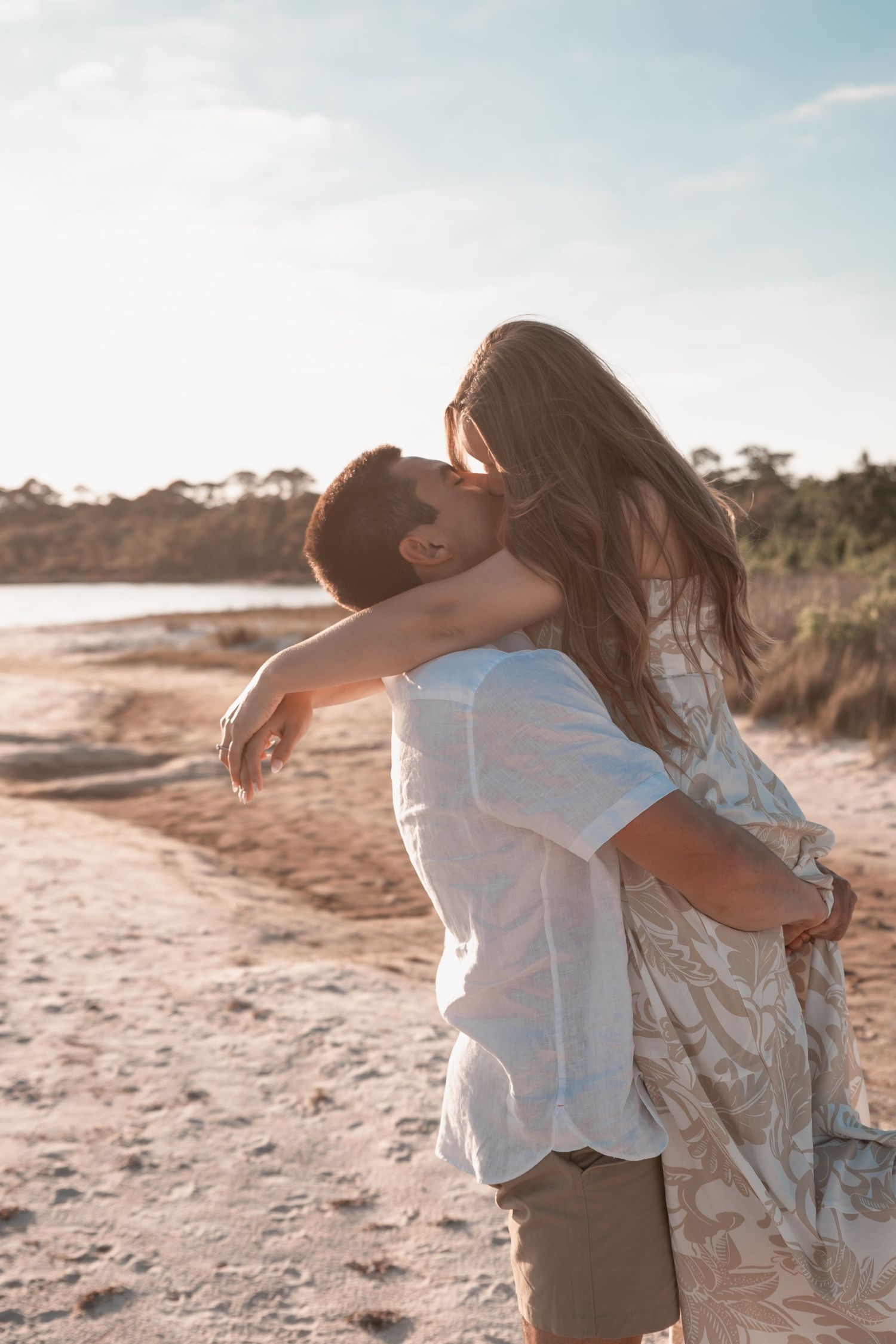 Couple shares a romantic kiss on a sandy beach at golden hour, woman wears a floral maxi dress.