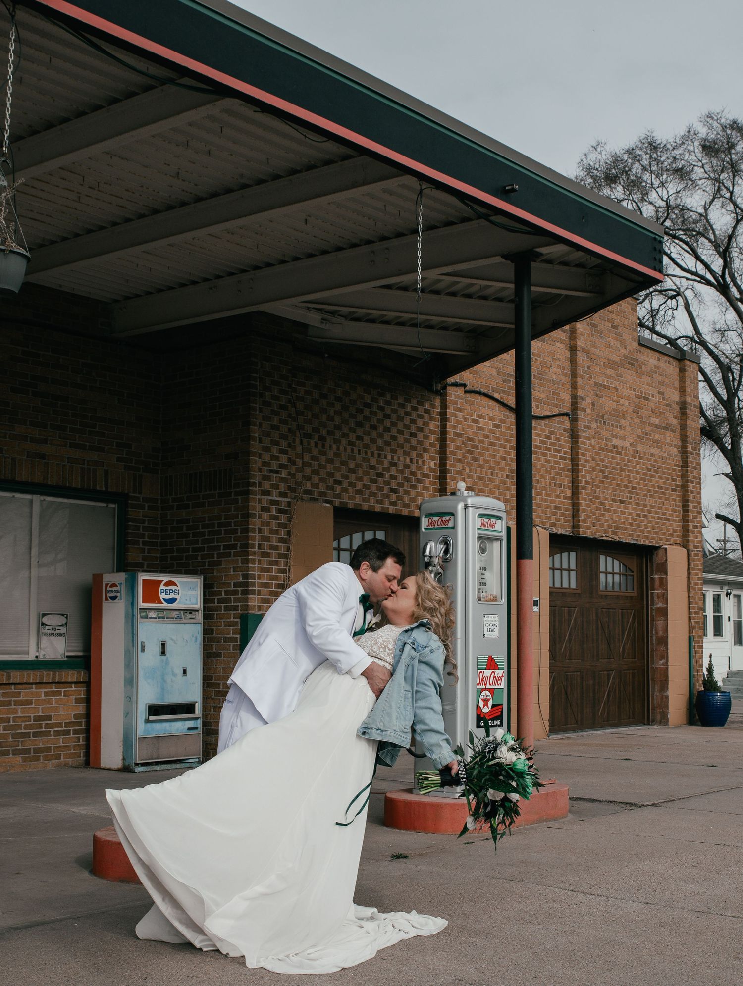 A bride and groom share a romantic dip kiss in front of a vintage brick gas station.