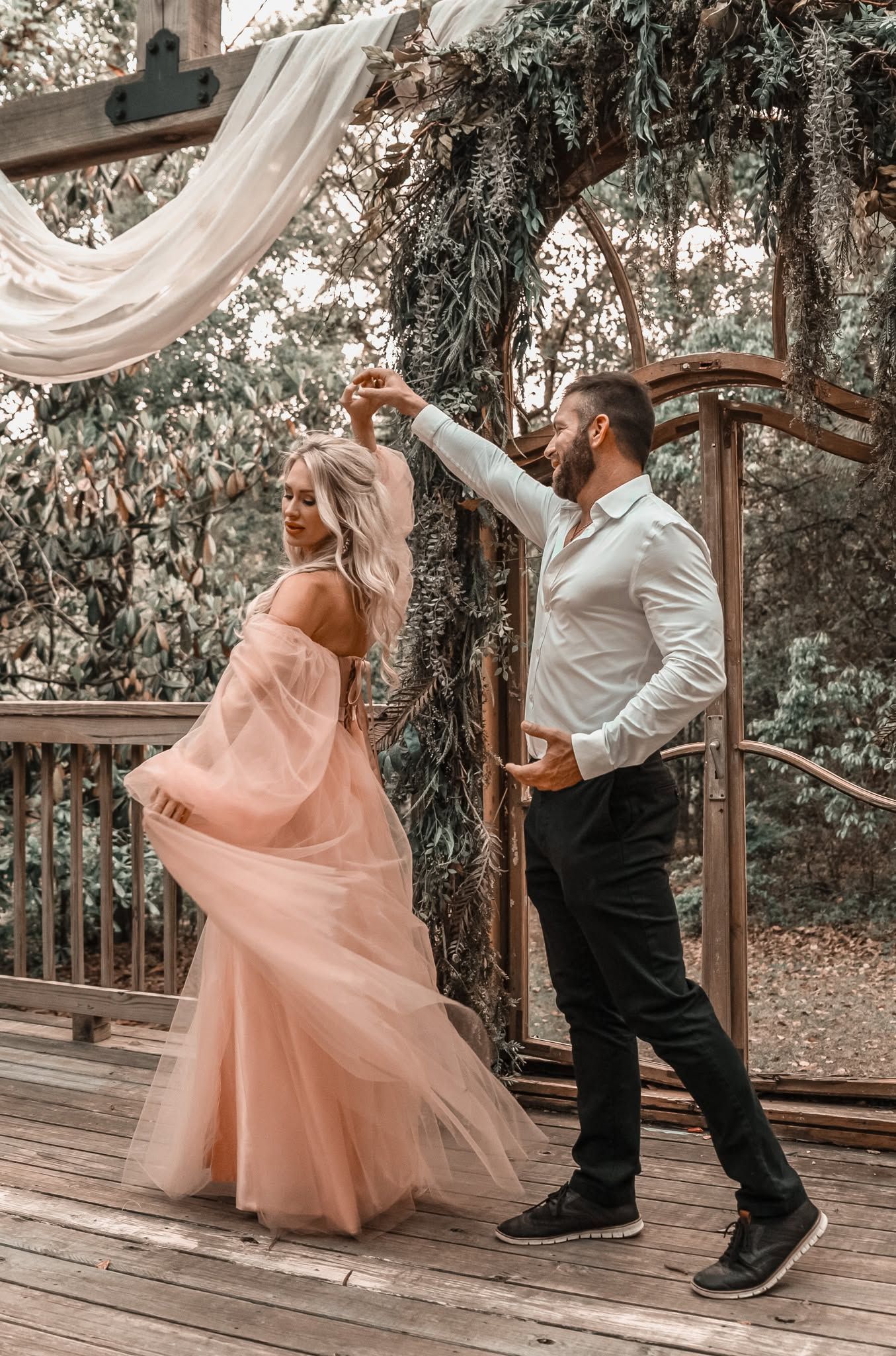 Couple dancing under a floral arch on a wooden deck, woman in pink tulle gown, man in white shirt.