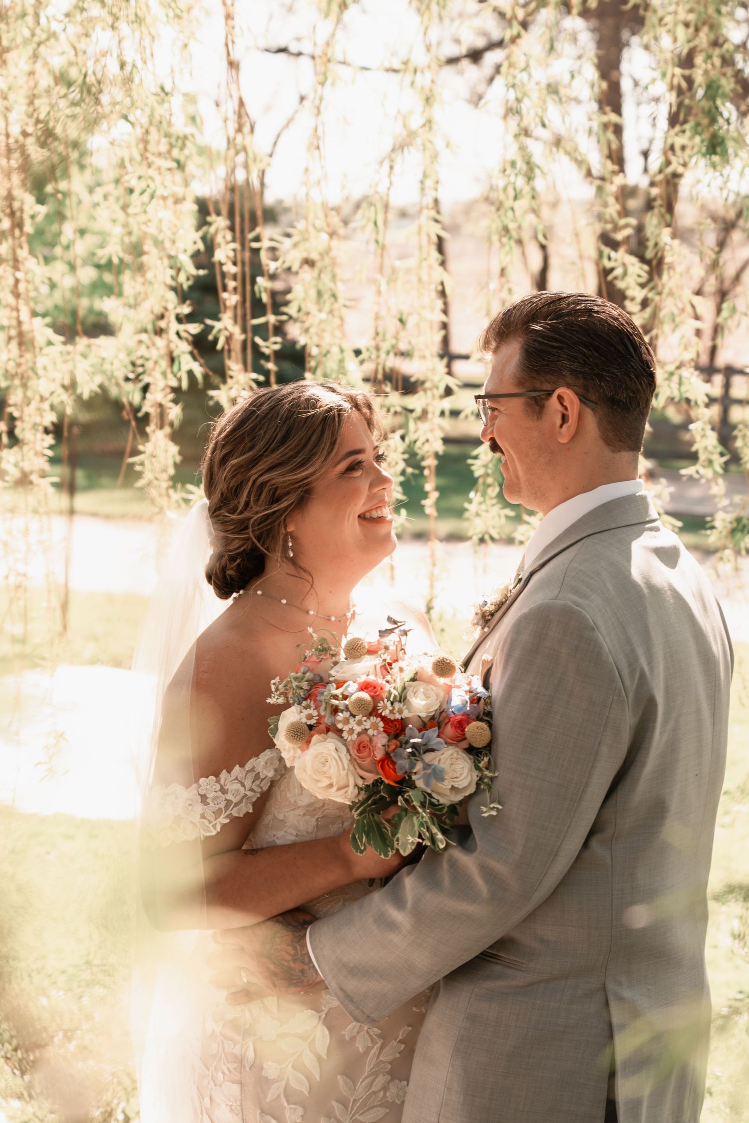 A bride and groom share a joyful moment under a weeping willow tree, bride holding a colorful bouquet.