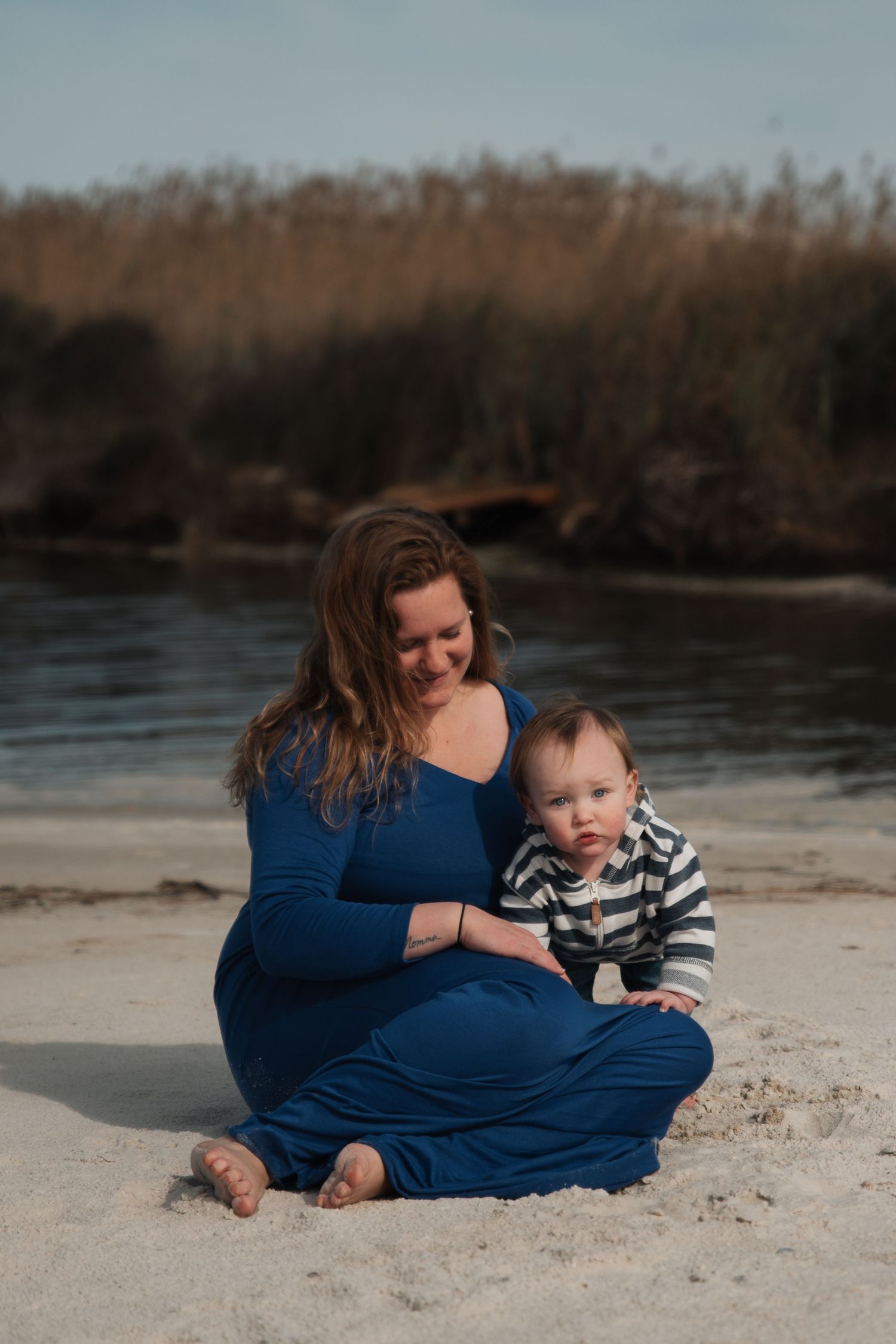 Pregnant woman in blue dress sits on sandy beach smiling at baby in striped outfit.