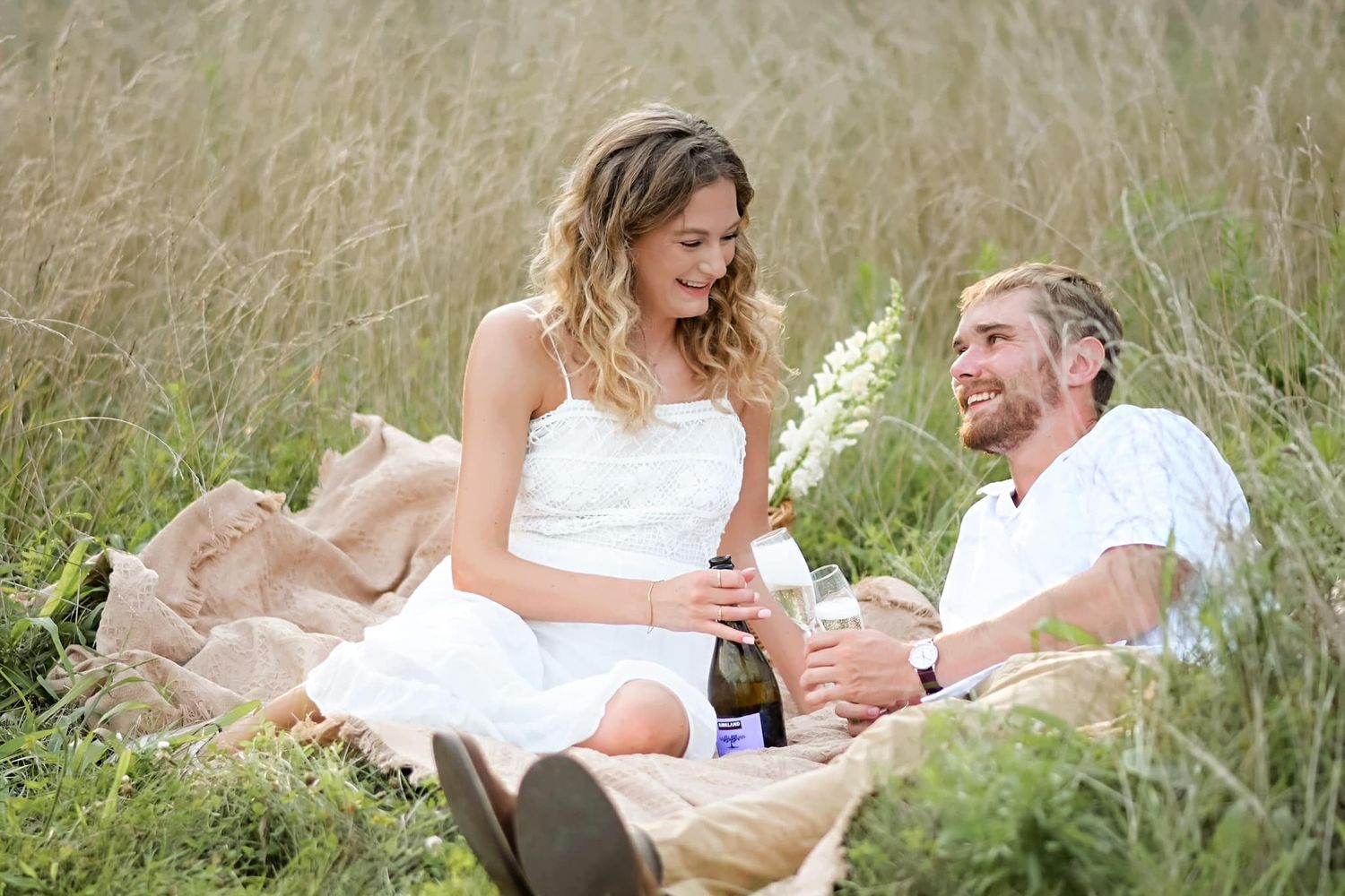 Couple relaxing in tall golden grass, sharing a bottle of champagne on a burlap blanket outdoors.