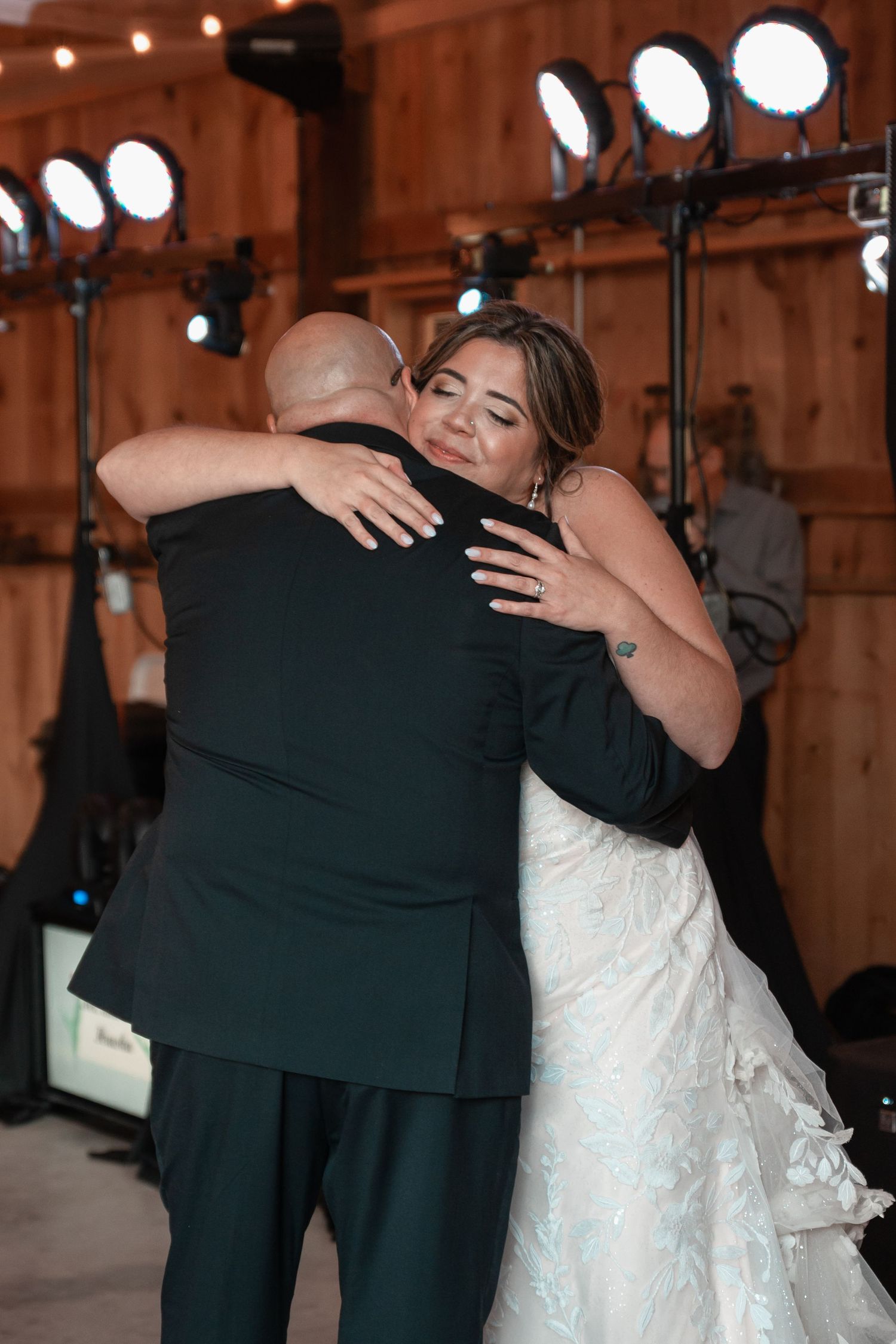 Bride in lace gown shares emotional hug with guest in barn wedding reception venue.