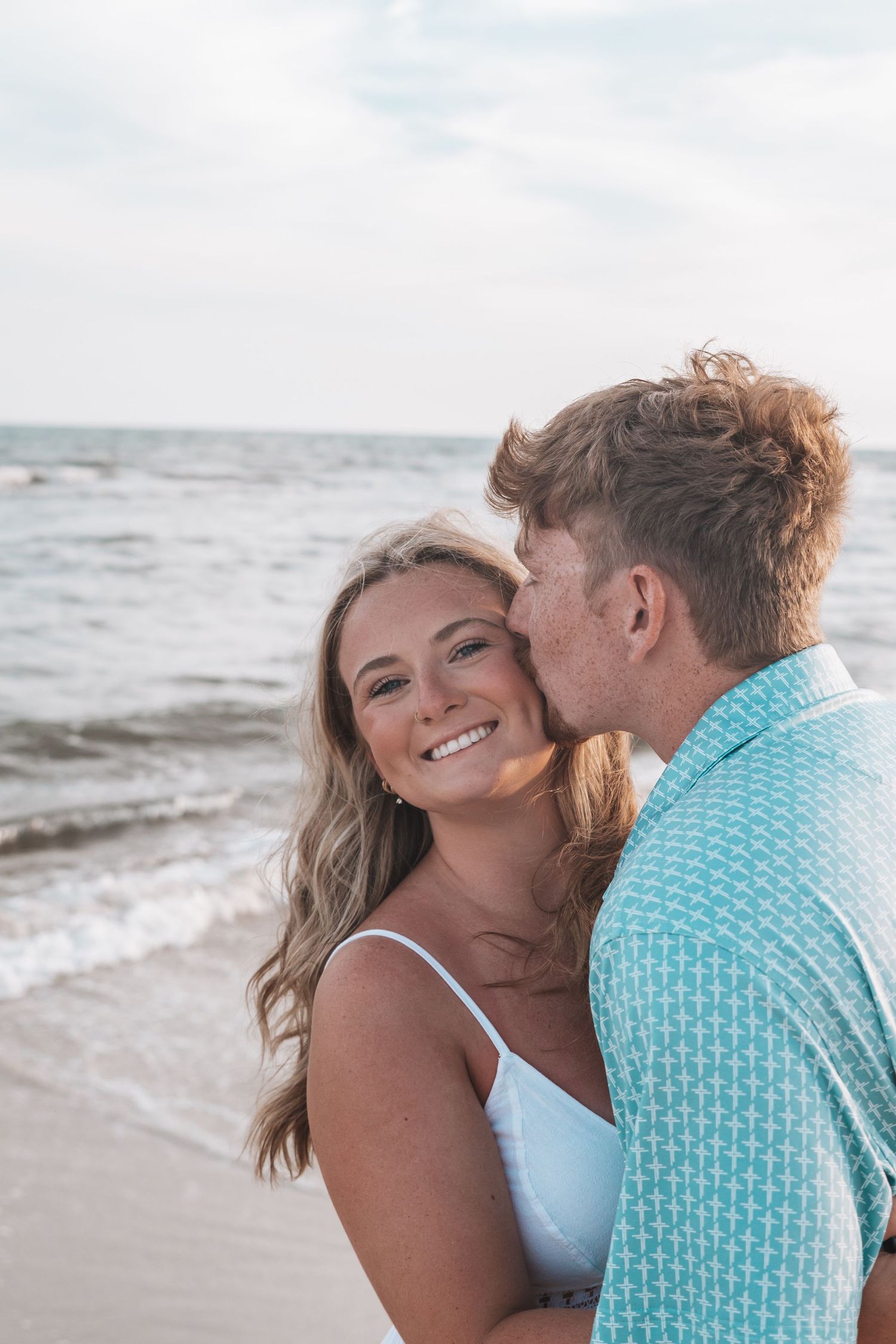A smiling woman in a white dress receives a kiss on the cheek from a man on a beach.